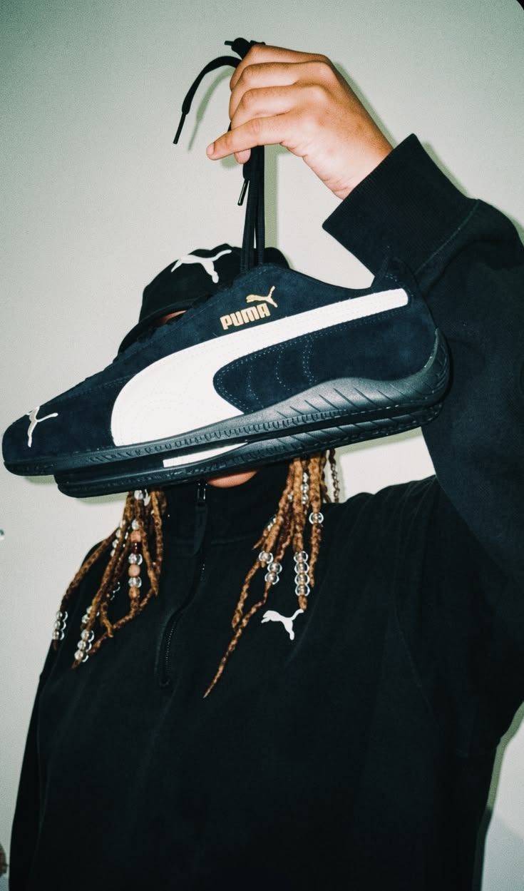 PUMA campaign image: a figure in a black PUMA zip-up and cap holding a navy suede PUMA sneaker with a white Formstrip by its laces in front of their face, beaded braids hanging below, shot with flash against a pale wall