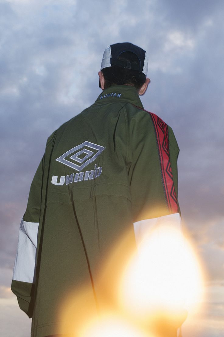 Umbro campaign image: a man in a black cap and an olive Umbro track jacket with the double diamond logo on the back and red taped sleeves, shot from behind against a cloudy sunset sky with lens flare