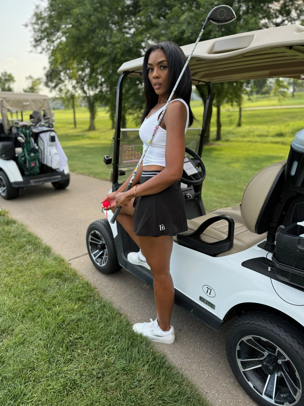Woman on a golf course beside a golf cart in a white cropped tank and a black skirt with a striped waistband and FIT BRIT logo, a driver over her shoulder