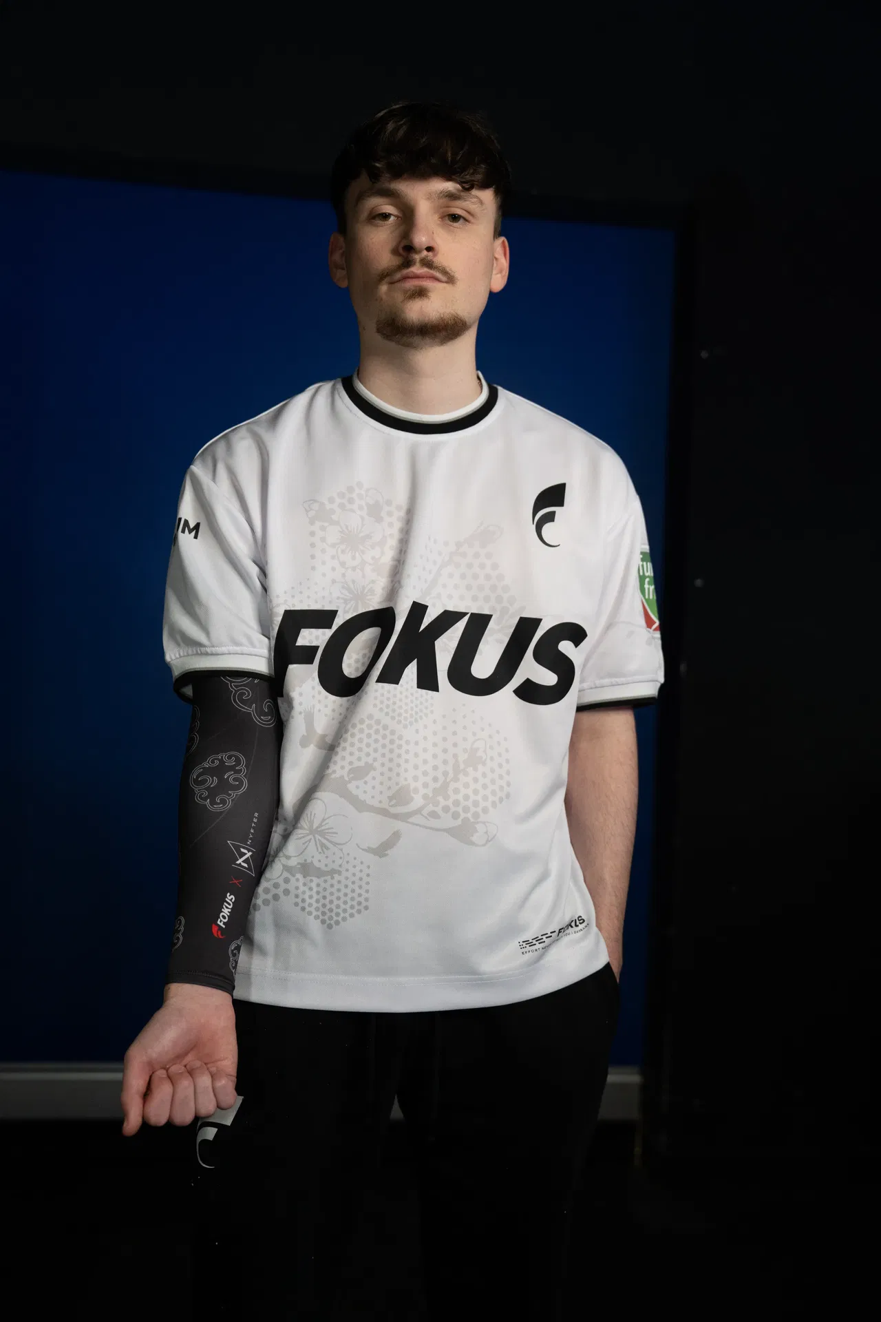 Young man wearing a white "Fokus" sports jersey and black sleeve against a dark blue and black background.