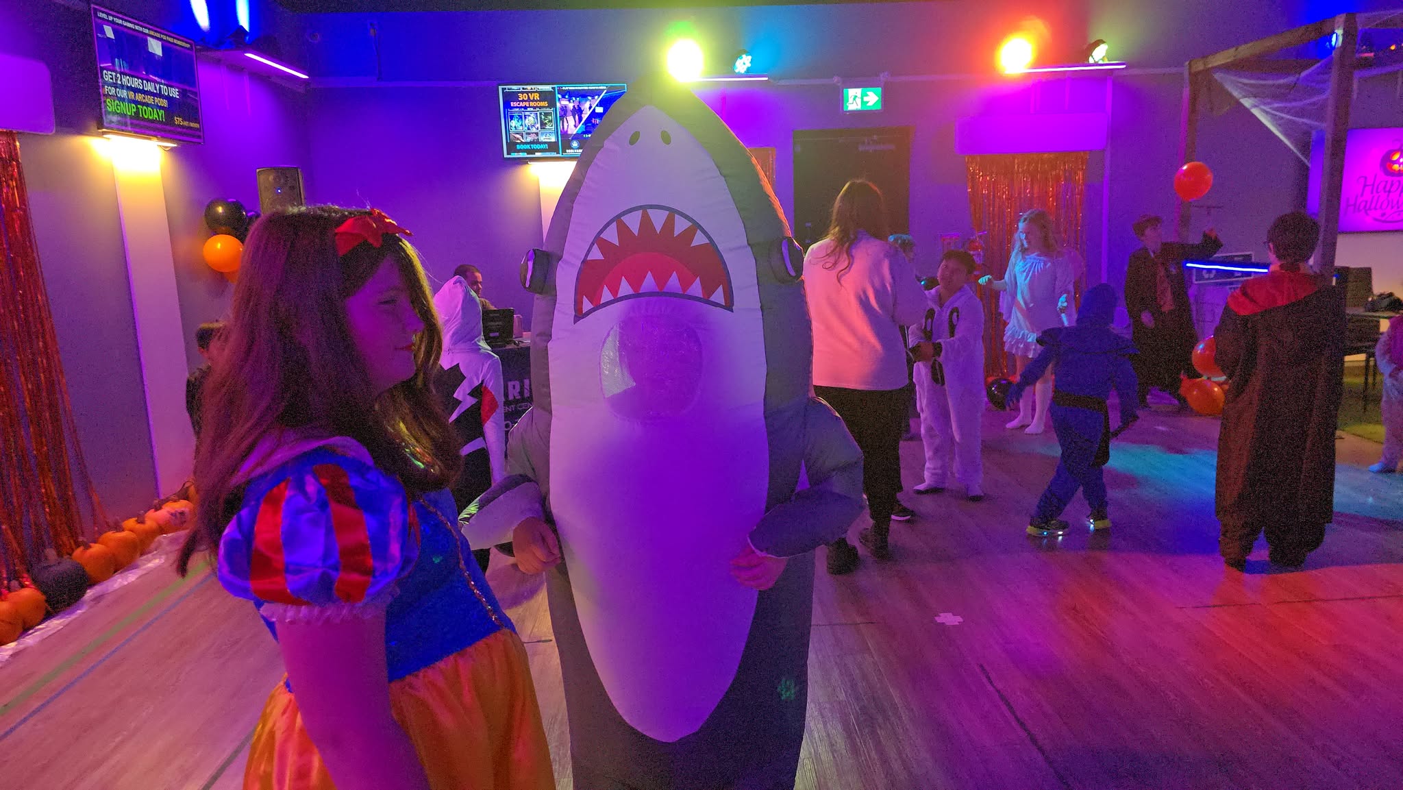 Kids in Halloween costumes, including Snow White and an inflatable shark, dancing under purple and orange party lights at Soulvaria