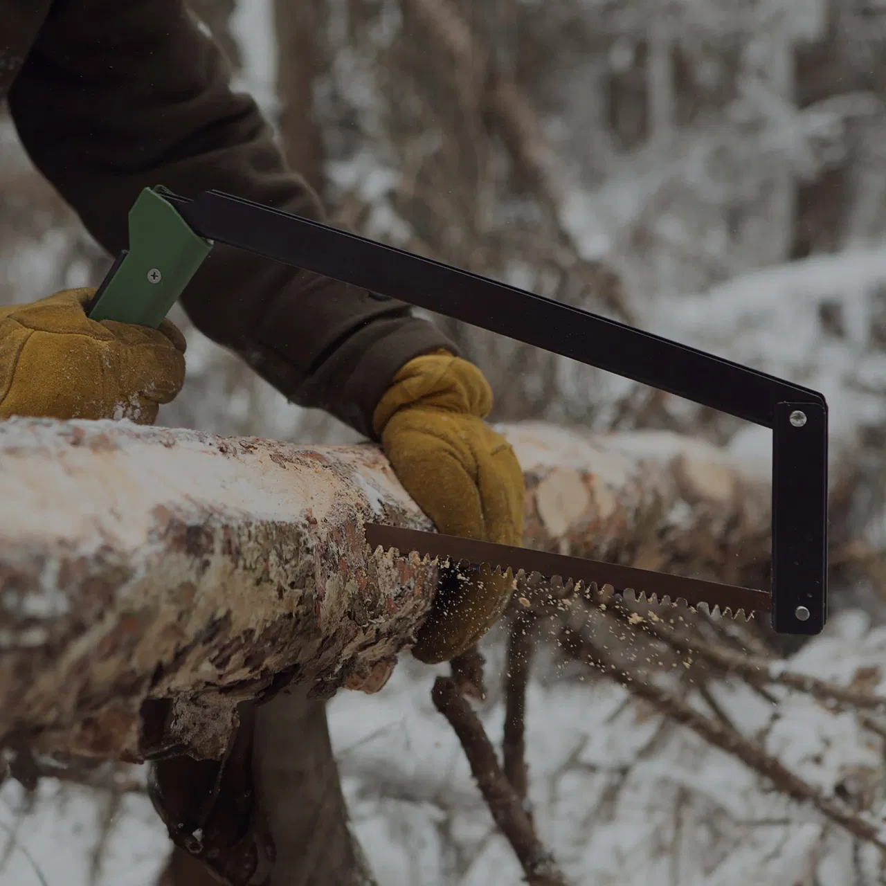 Person sawing a snow-covered log with a green-handled bow saw, wearing yellow gloves.