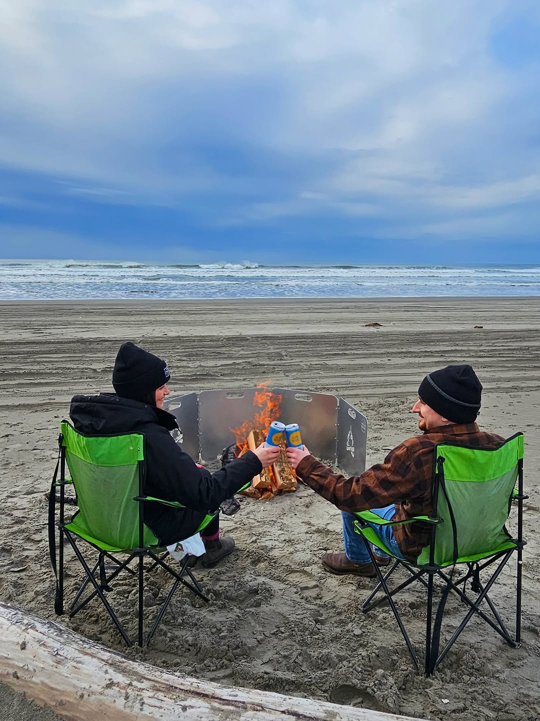 Two people sit in green camp chairs on a sandy beach, clinking blue cans in a toast over a campfire shielded by the Original Fire Reflector.