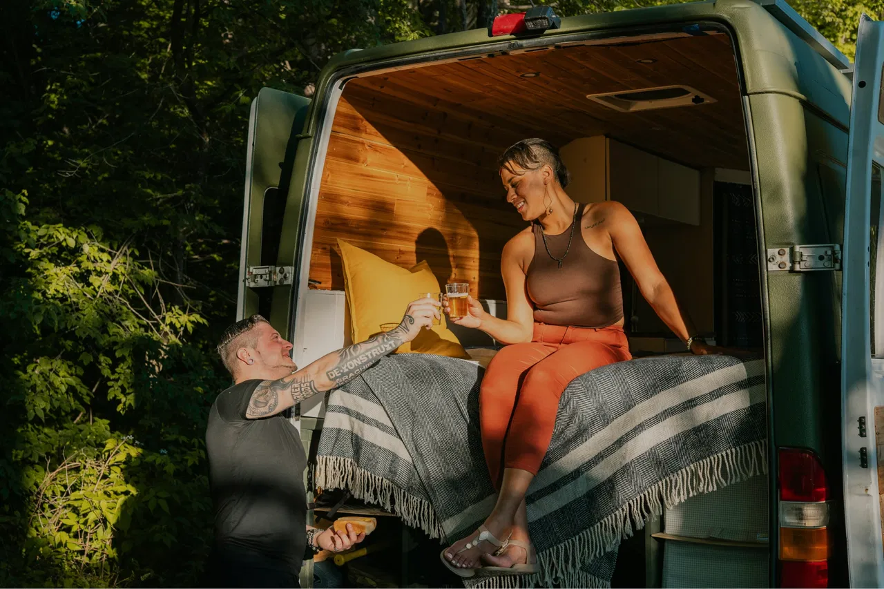 A couple enjoys a drink at the back of a camper van in a wooded area.