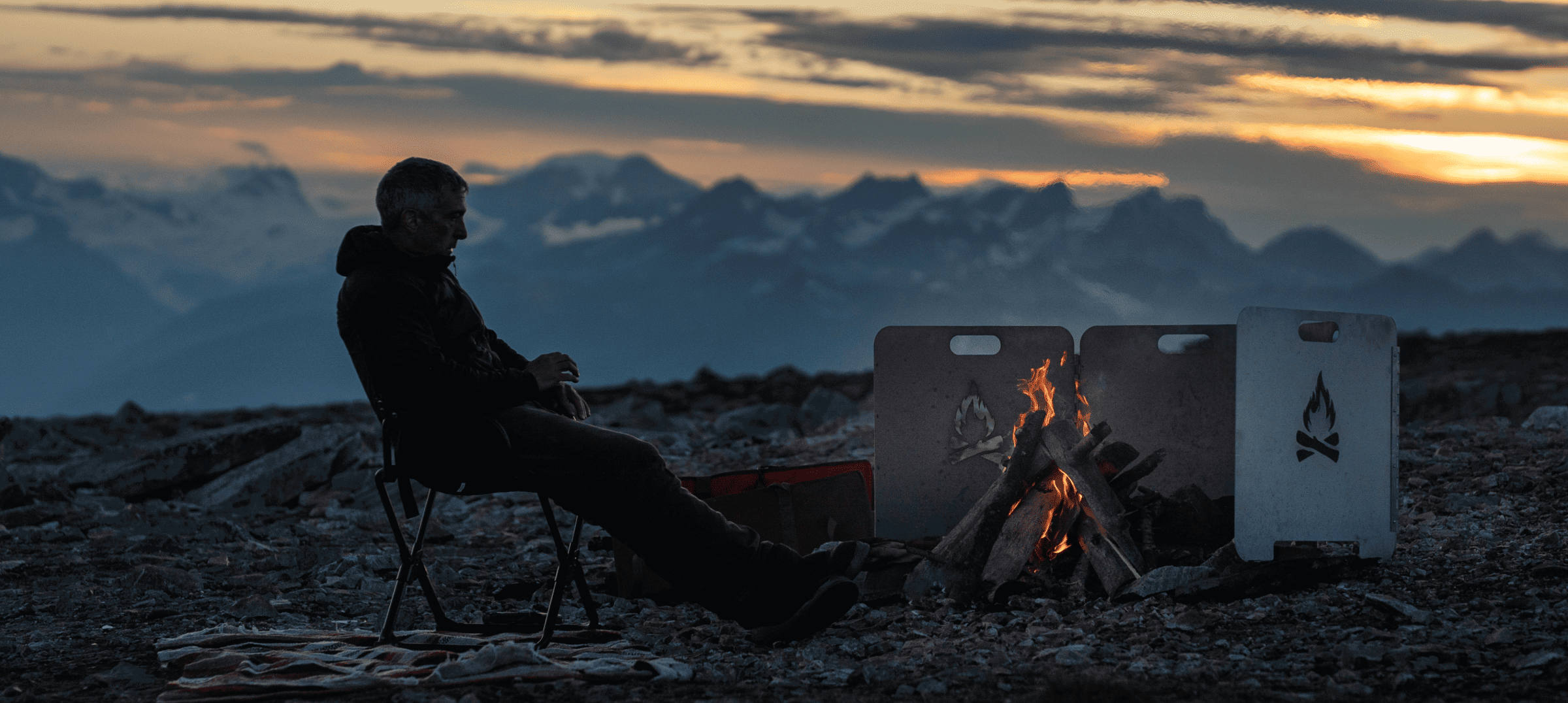 Person sitting by a campfire on rocky terrain at sunset with mountains in the distance.