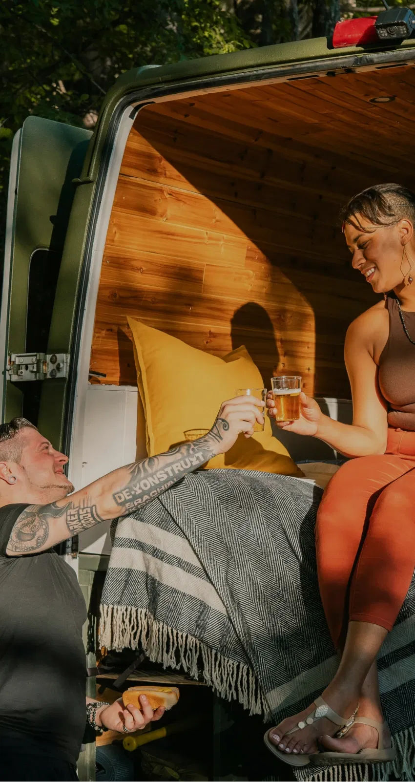 A couple enjoys a drink at the back of a camper van in a wooded area.