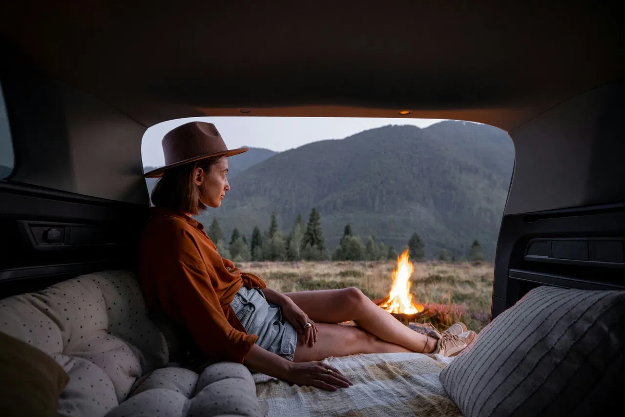 Woman in car trunk watching campfire, mountains in background.