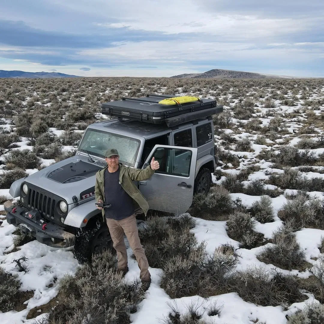 Man giving thumbs up next to a gray Jeep in a snowy, shrub-filled landscape.