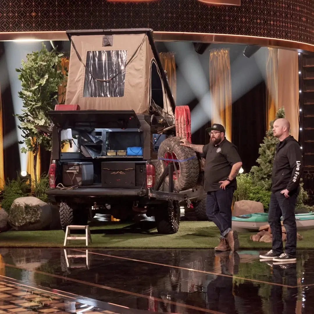 Two men stand by a rugged off-road vehicle with an open rooftop tent and gear on display.
