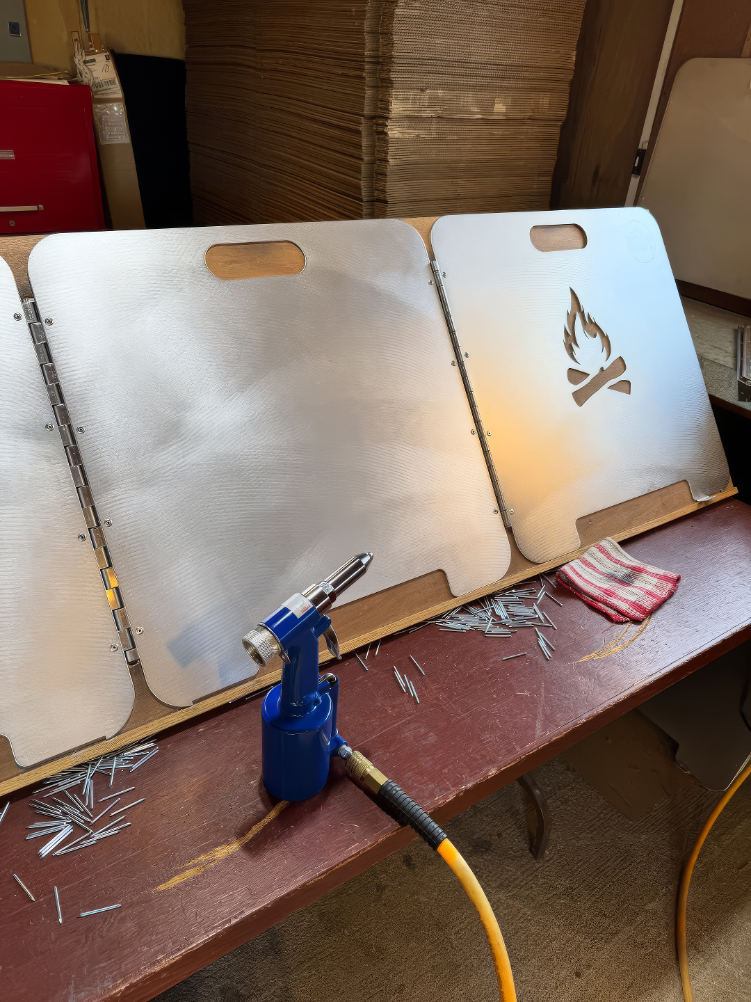 Close-up of the Original Fire Reflector panels being assembled on a workshop table next to a blue rivet gun and metal rivets.