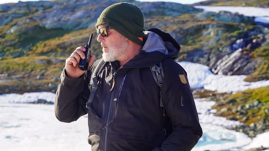 Man wearing a green beanie and sunglasses using a walkie-talkie in a snowy mountainous area.