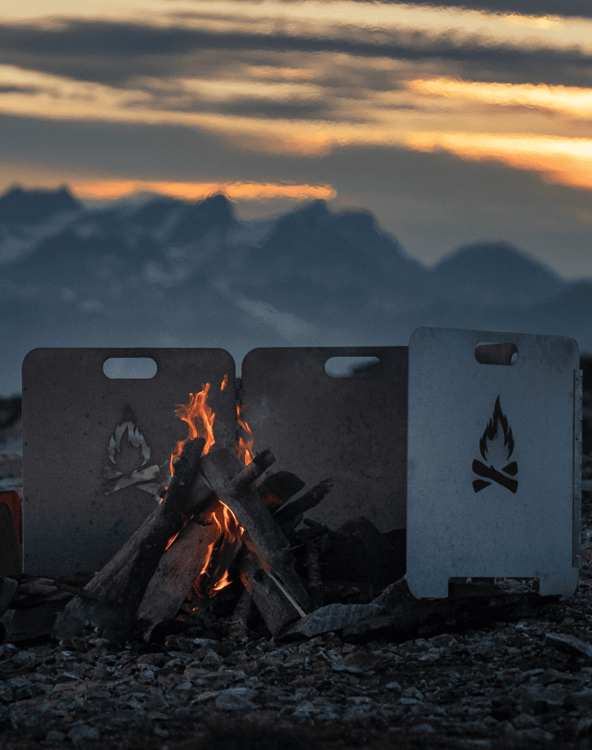 Person sitting by a campfire on rocky terrain at sunset with mountains in the distance.