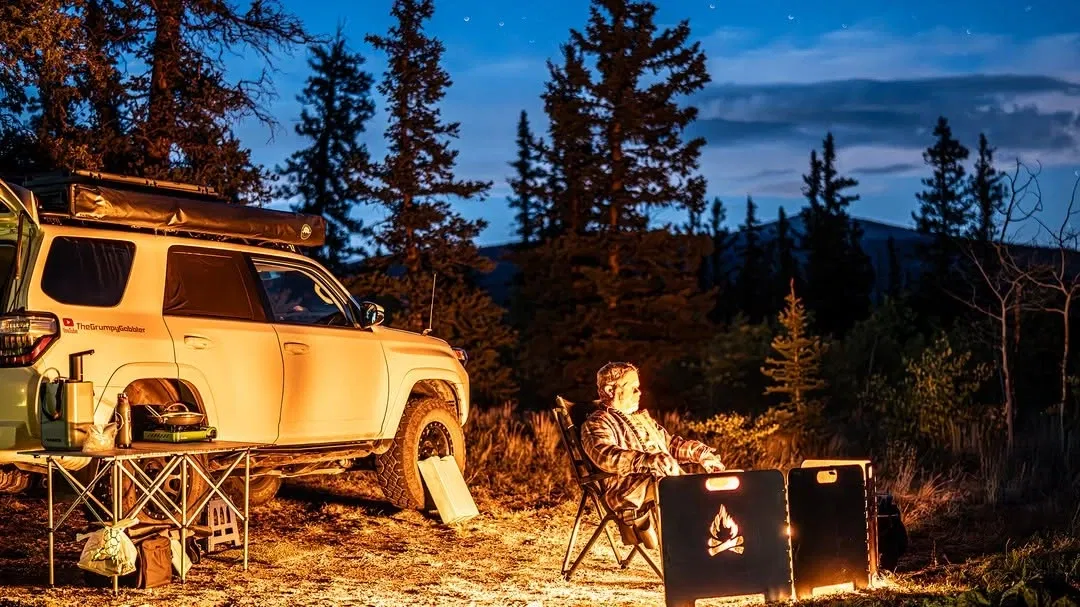 Person sitting by a campfire next to an SUV in a forest at dusk.
