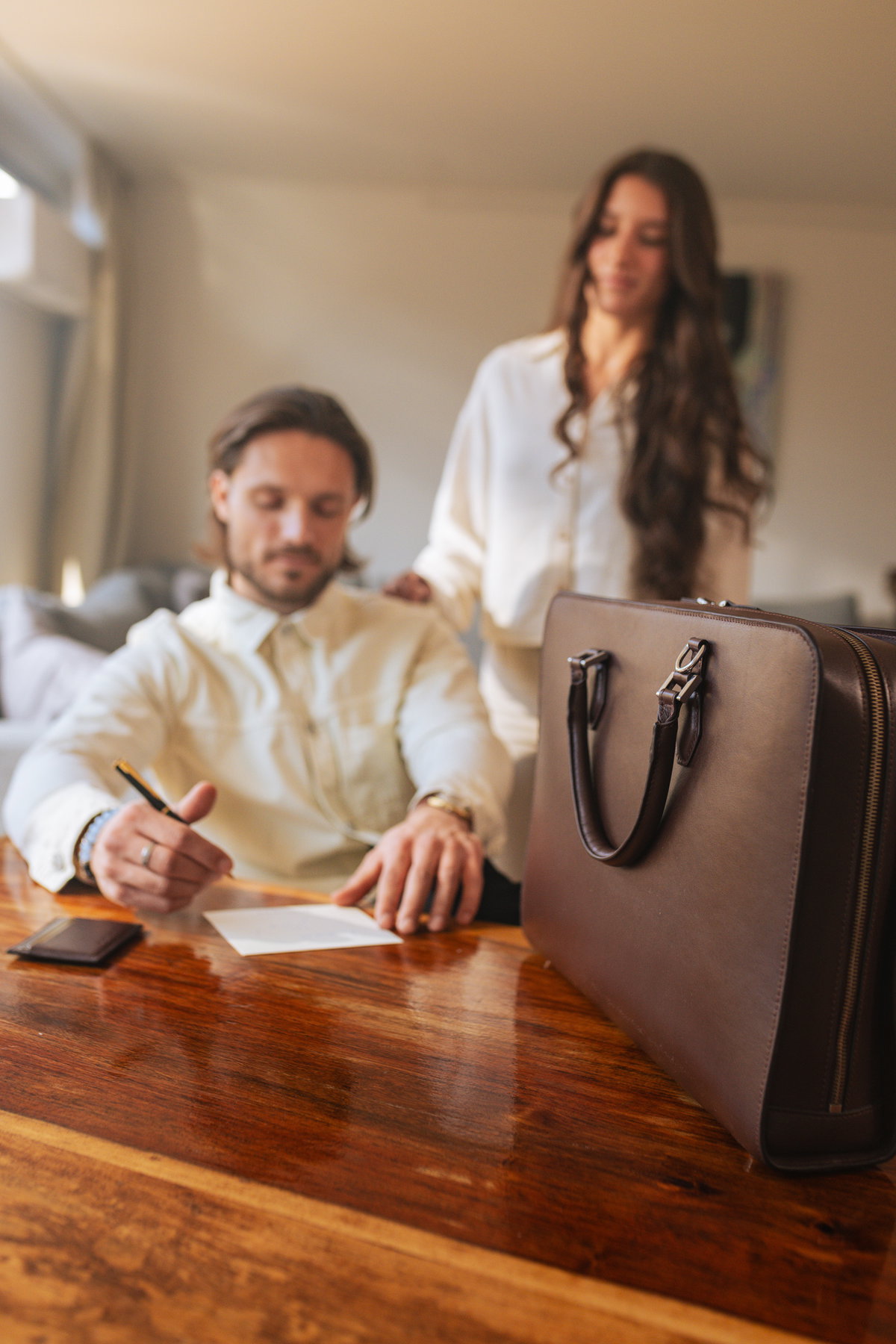 Man writing at a wooden desk beside a dark brown leather briefcase