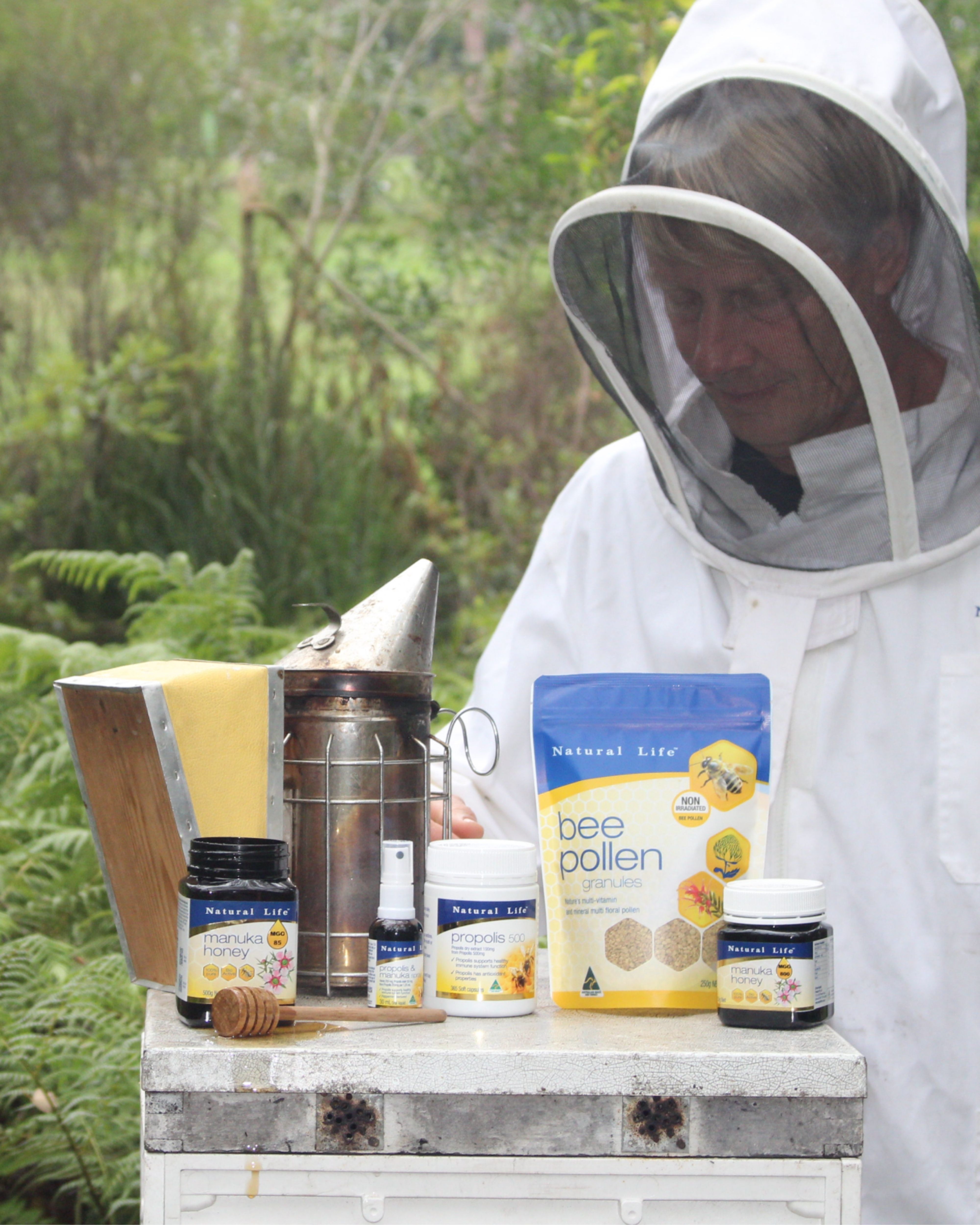 Beekeeper beside a hive with Natural Life Manuka honey, propolis, bee pollen and smoker in front of Australian ferns