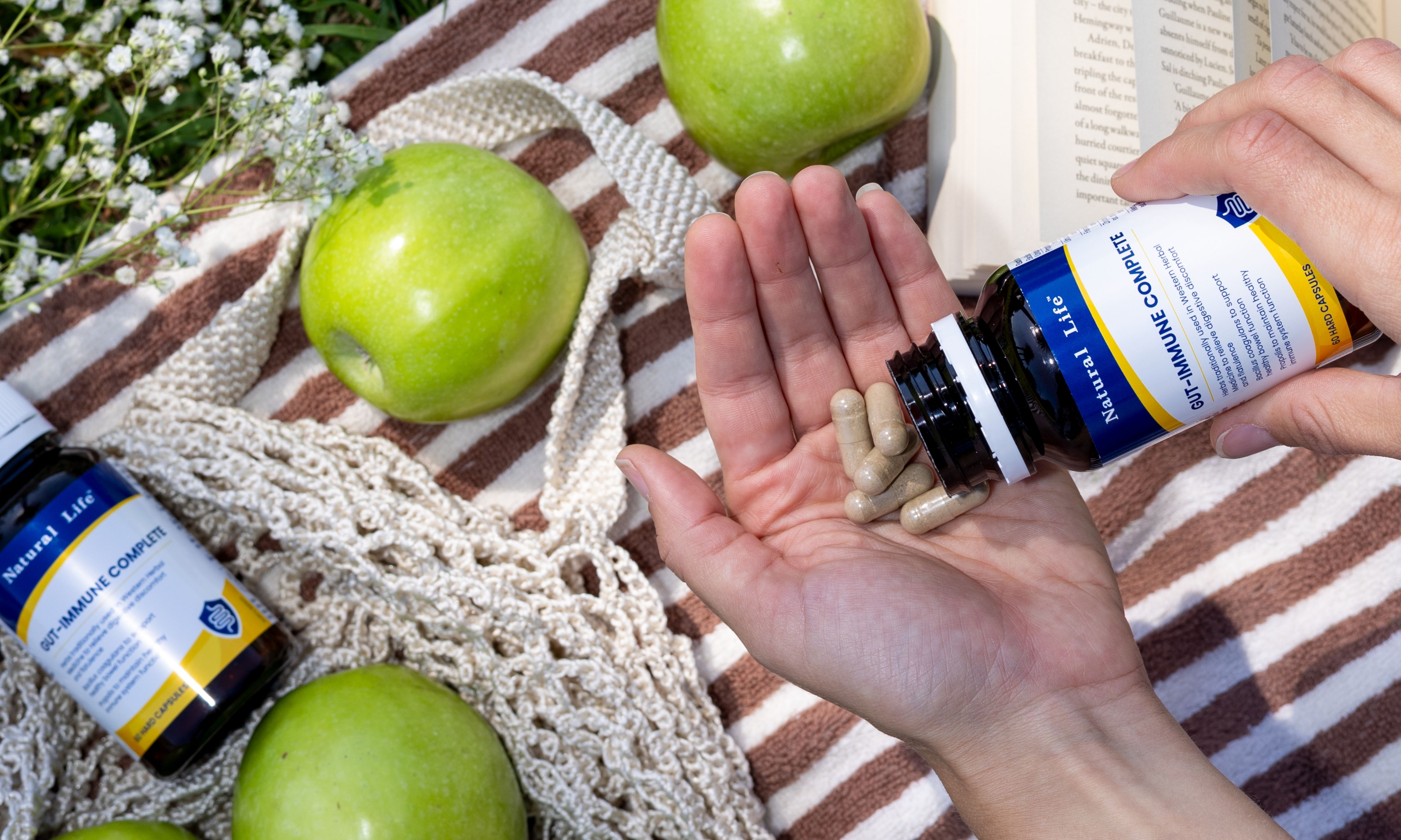 Hands tipping Gut-Immune Complete capsules from the bottle onto an open palm, on a picnic blanket with green apples, a string bag and a book