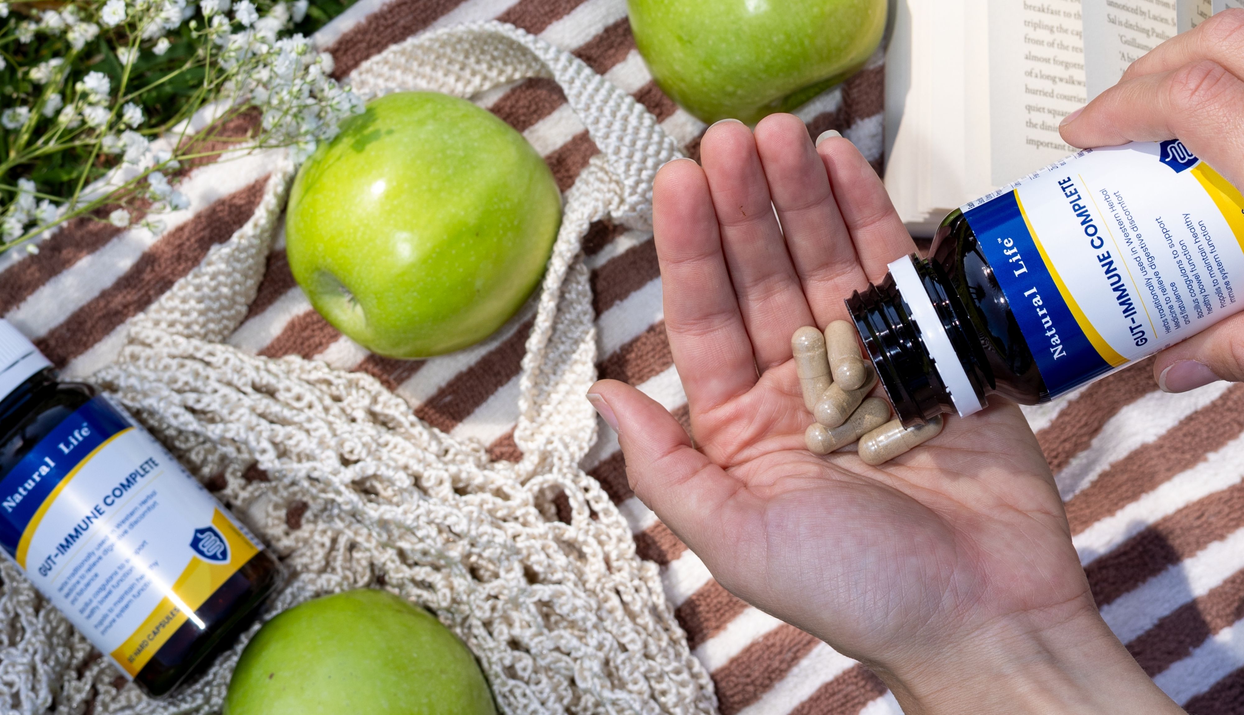 Gut-Immune Complete capsules poured into an open hand from the bottle, on a striped picnic blanket with green apples