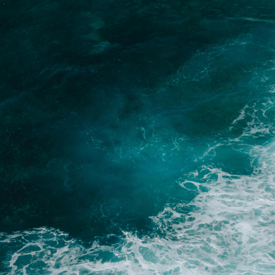 Deep teal ocean water with white surf breaking across the surface, seen from above