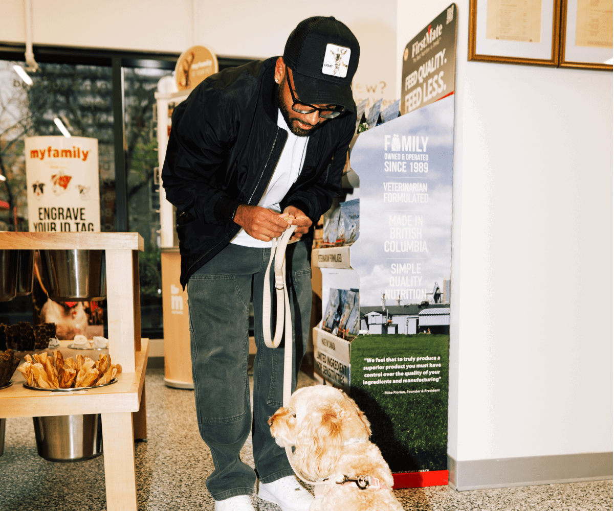 A customer giving a treat to their fluffy dog inside Pet Planet, with a FirstMate brand display banner in the background.