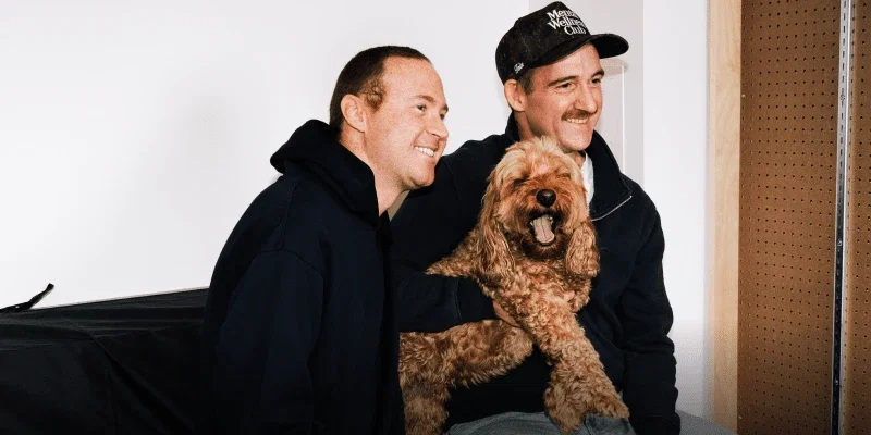 Two friends laughing together while holding a happy goldendoodle on their lap