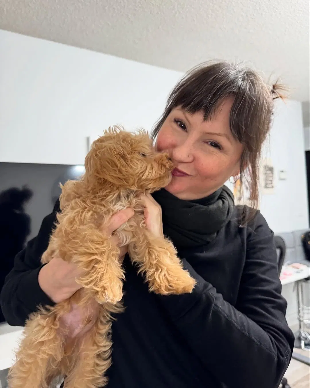 A woman smiling while holding and kissing a small curly-haired brown puppy indoors