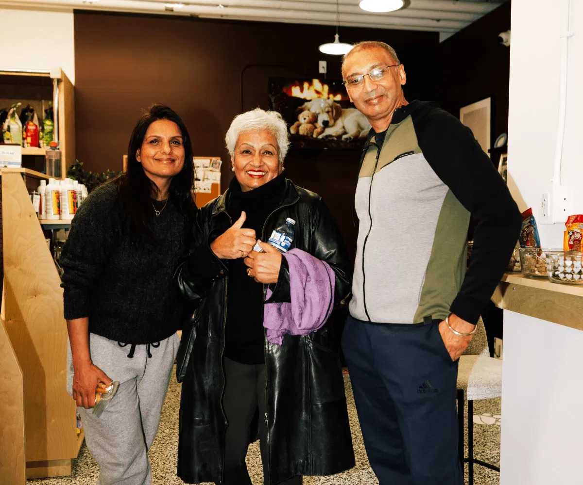 Three smiling customers pose together inside Pet Planet Toronto. The woman in the center gives a thumbs up, wearing a black leather jacket, with pet product shelves and a puppy photo visible in the background.