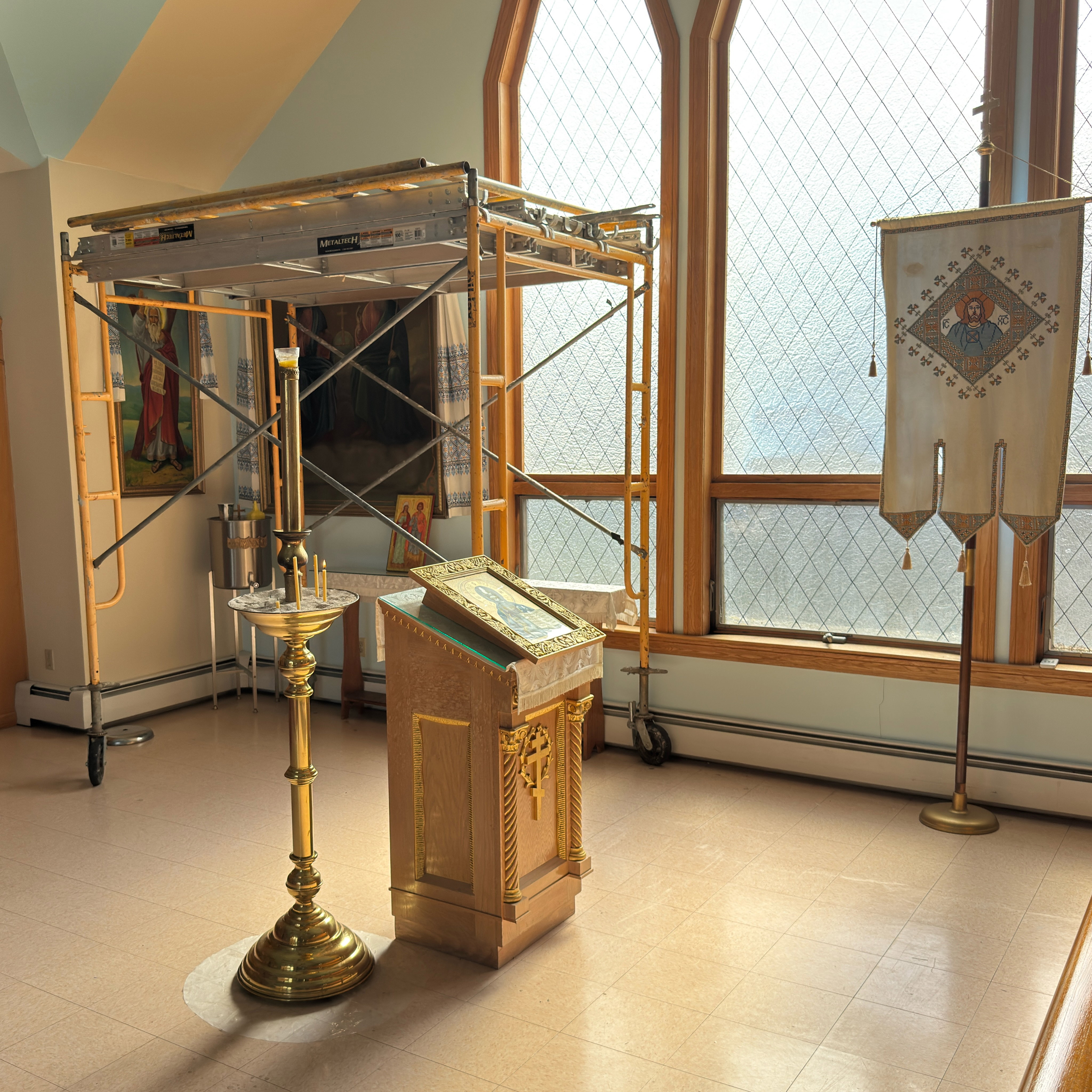 A sunlit corner of Holy Trinity during the icon work: painters' scaffolding stands before wall icons of saints hung with embroidered rushnyky, beside tall leaded-glass arched windows; in front, a brass candle stand with thin beeswax tapers, a carved golden analogion holding a framed icon, and an embroidered church banner bearing the face of Christ