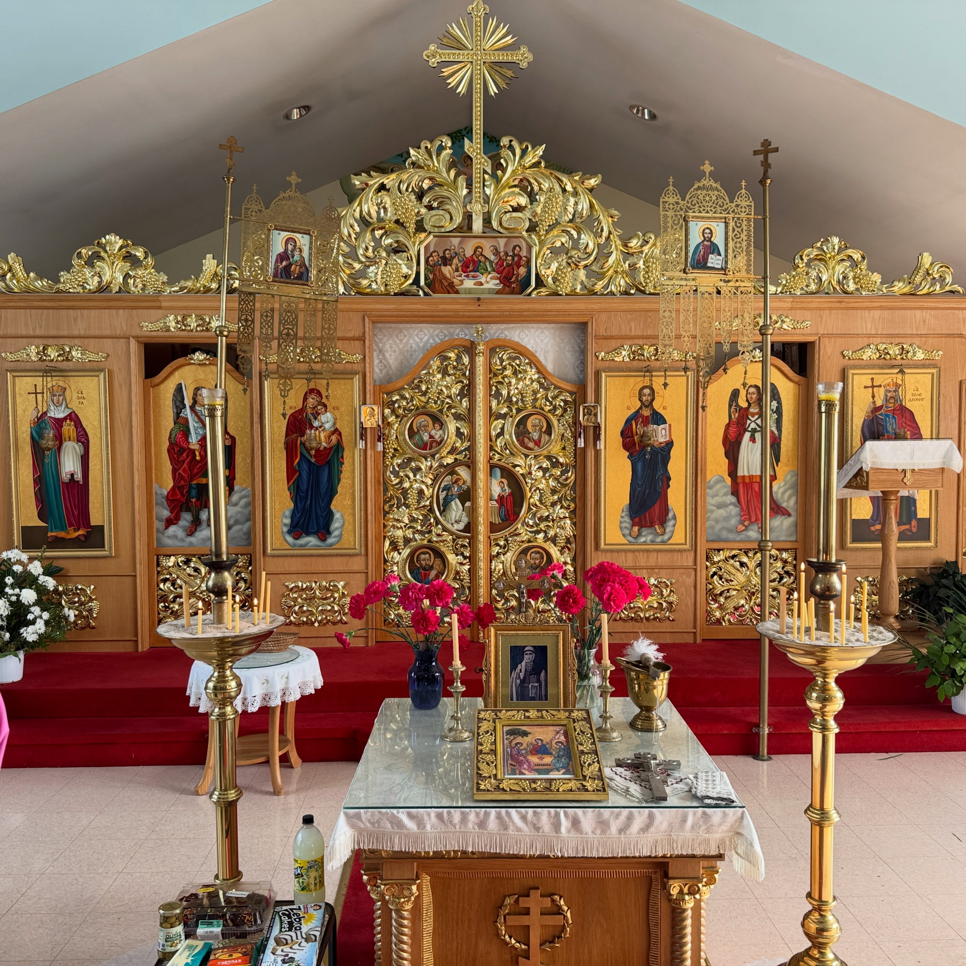 The gilded iconostasis at Holy Trinity: carved gold Royal Doors with round icons of the Annunciation and the Evangelists, flanked by icons of St. Olga, Archangel Michael, the Theotokos with the Christ Child, Christ, Archangel Gabriel and St. Volodymyr, beneath the Last Supper and a radiant gold cross; in front, a table with a Holy Trinity icon, beeswax candles and red carnations