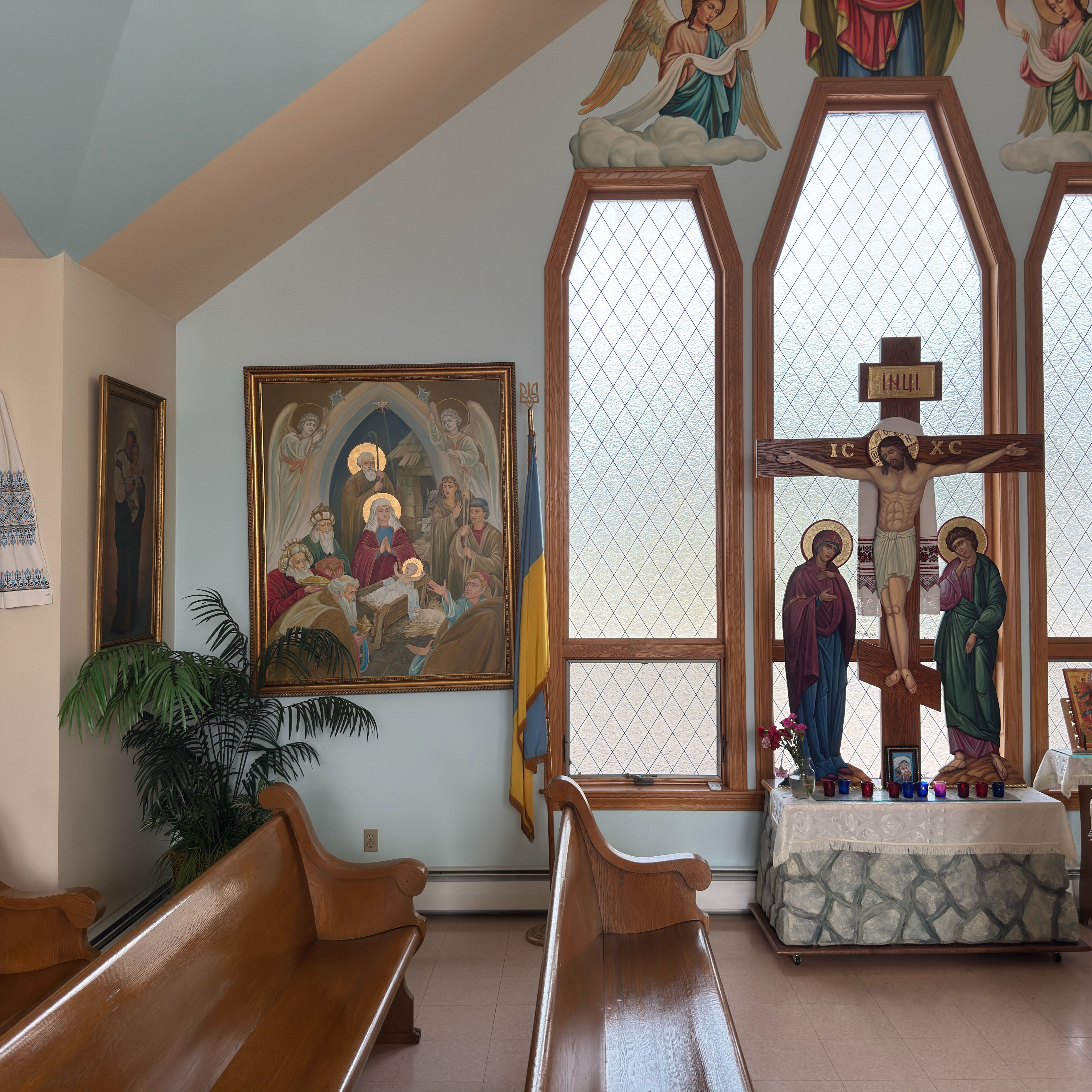 A bright corner of Holy Trinity beneath the sloped ceiling: a large gilt-framed painting of the Nativity of Christ shows the Christ Child in the manger with the Theotokos and St. Joseph, angels, the Magi bearing gifts and the shepherds; beside it a Ukrainian flag topped with a gold trident, and to the right the painted Crucifixion inscribed IC XC and INЦІ, draped with an embroidered rushnyk, with the Theotokos and St. John beneath the cross before tall leaded-glass windows, above a Golgotha table of votive candles, carnations and a small icon; angels holding a white cloth are painted above the windows, with a potted palm, wall icons, an embroidered rushnyk and polished wooden pews in front