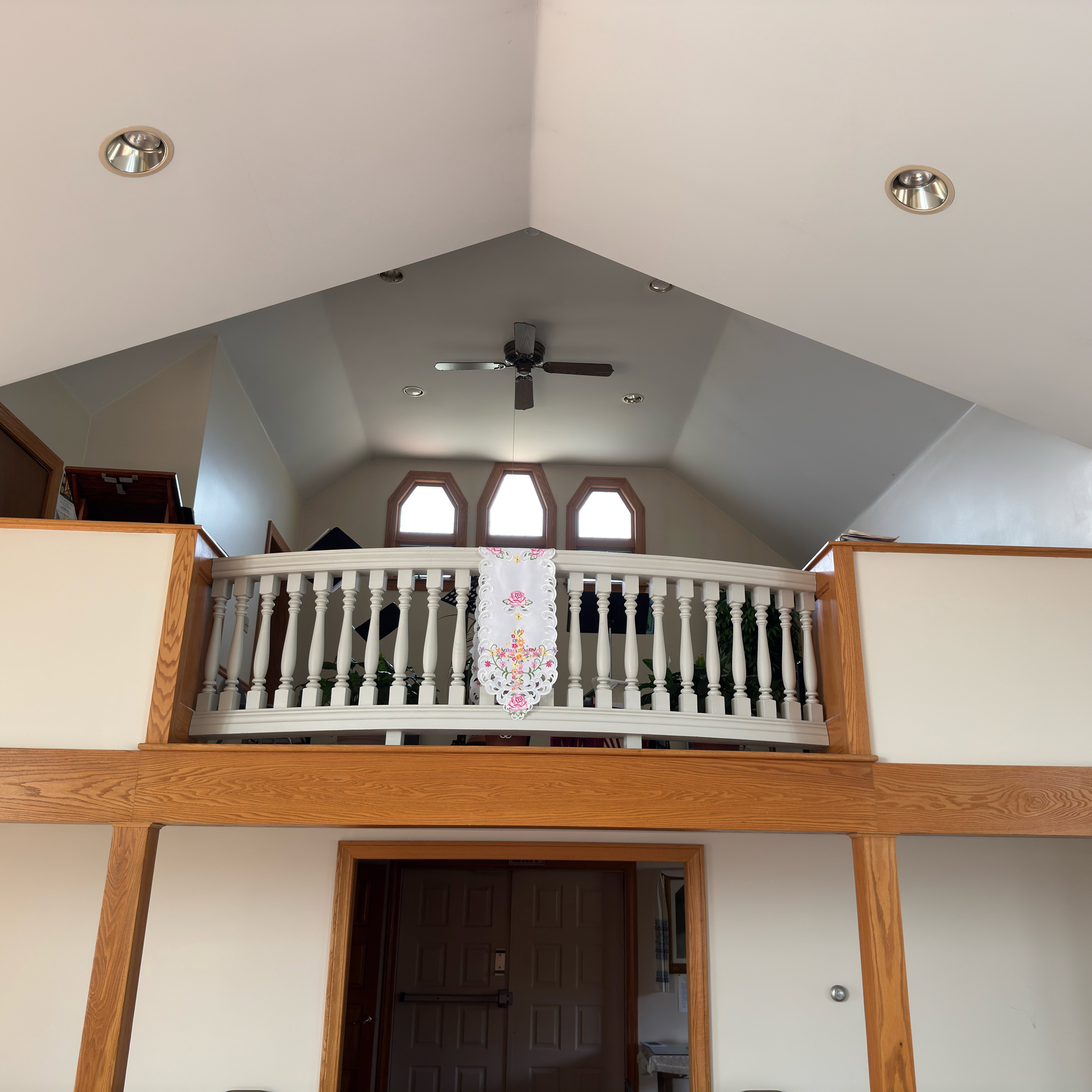 The choir loft at Holy Trinity, seen from the nave: a curved white balustrade draped with an embroidered cloth of pink roses and spring flowers, framed in golden oak beams above the church's main entrance doors; behind the rail stand music stands and green plants beneath three tall windows and a vaulted ceiling with a ceiling fan