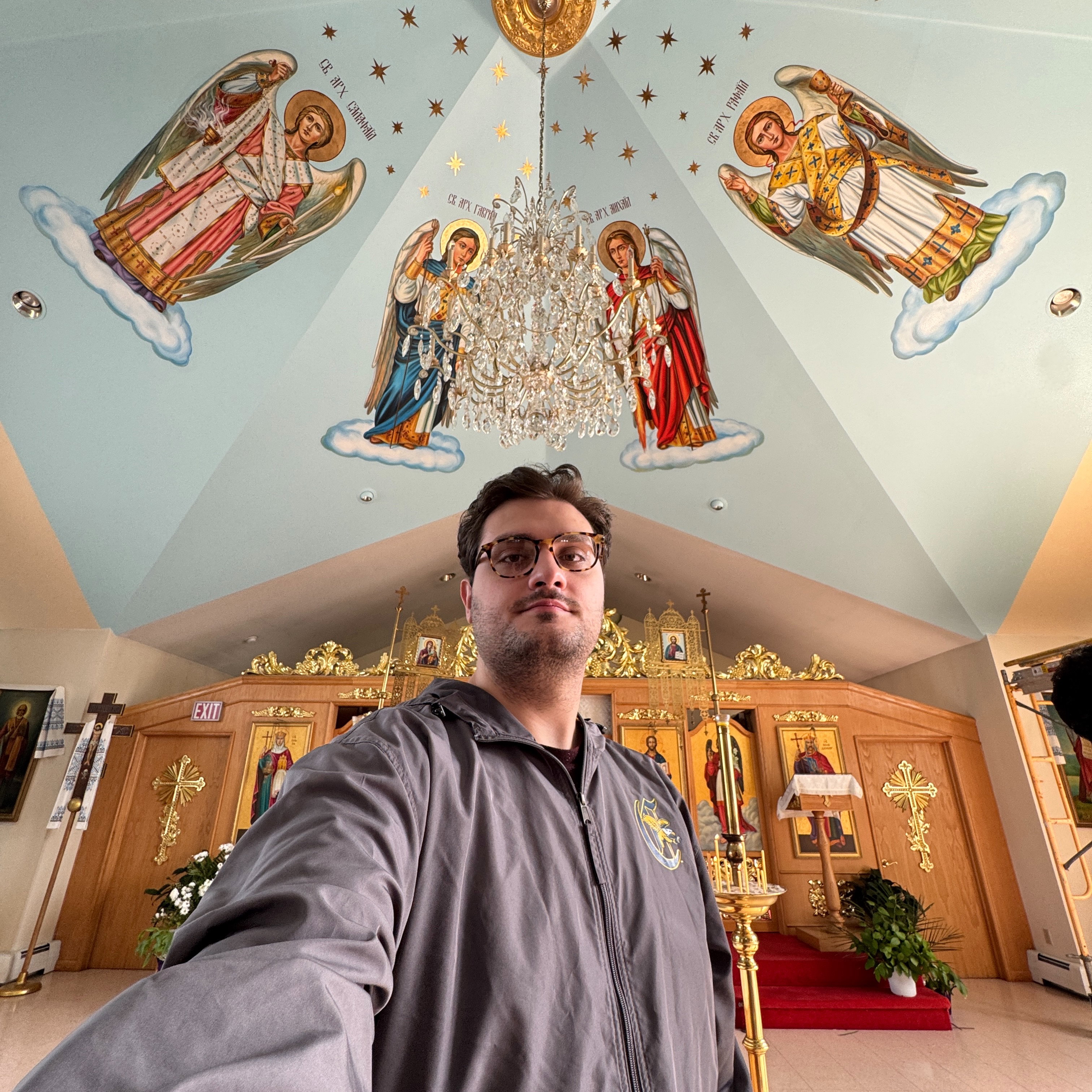 Professor Kristiyan-Daniel V. Iliev, in tortoiseshell glasses and a grey zip jacket, stands in the nave of Holy Trinity beneath the ceiling fresco of the archangels, each named in Church Slavonic (Cyrillic) lettering: Selaphiel with a censer, Gabriel in blue, Michael in red and Raphael in gold vestments, standing on clouds among gold stars around a crystal chandelier; behind him the gilded iconostasis with its icons, brass candle stands and red-carpeted steps
