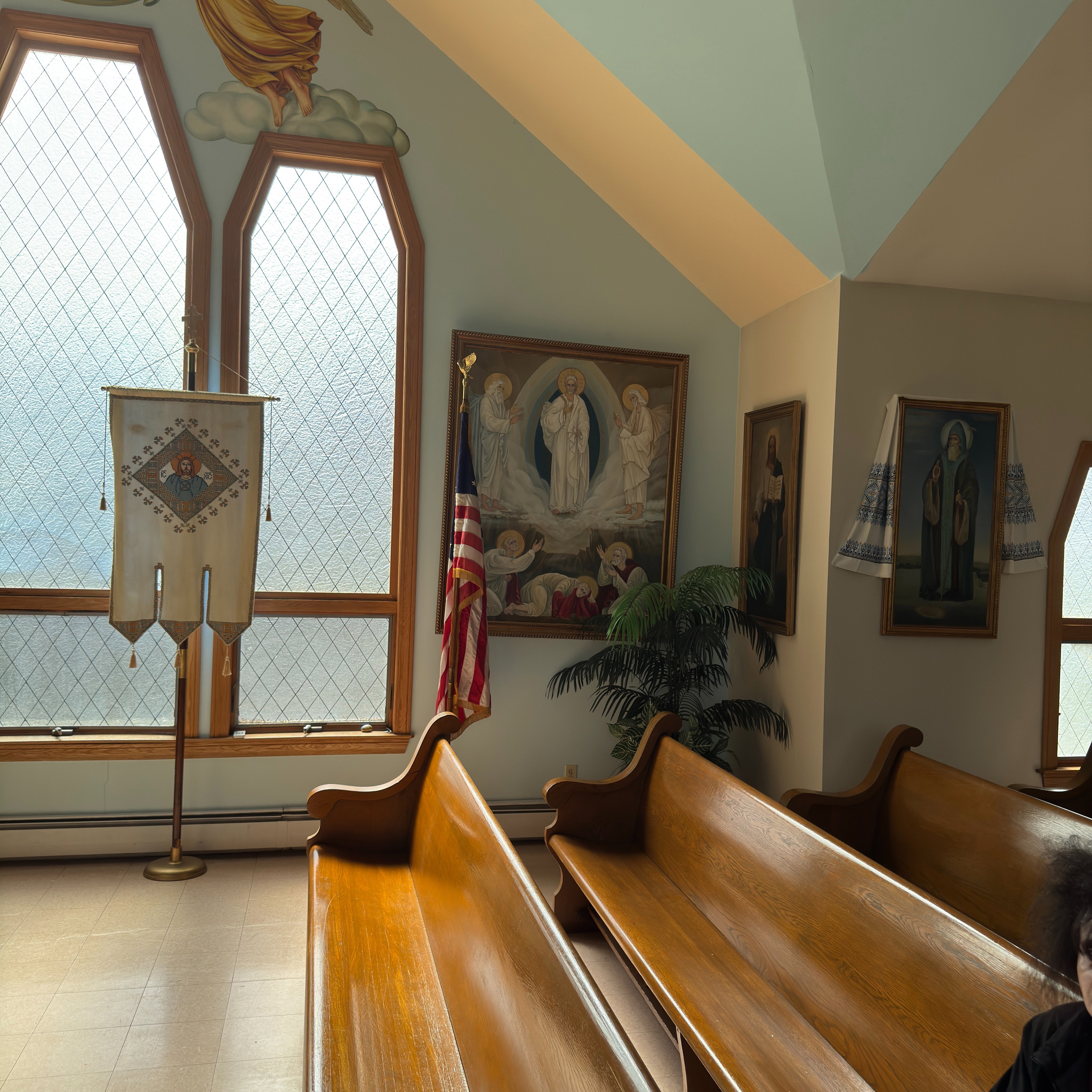 A sunlit side of the nave at Holy Trinity: a large gilt-framed painting of the Transfiguration shows Christ in white glory between Moses and Elijah above the three fallen apostles; beside it an American flag, a potted palm, icons of saints (one draped in an embroidered rushnyk) and an embroidered processional banner bearing the face of Christ before tall leaded-glass windows, beneath an angel fresco, with rows of polished wooden pews in front