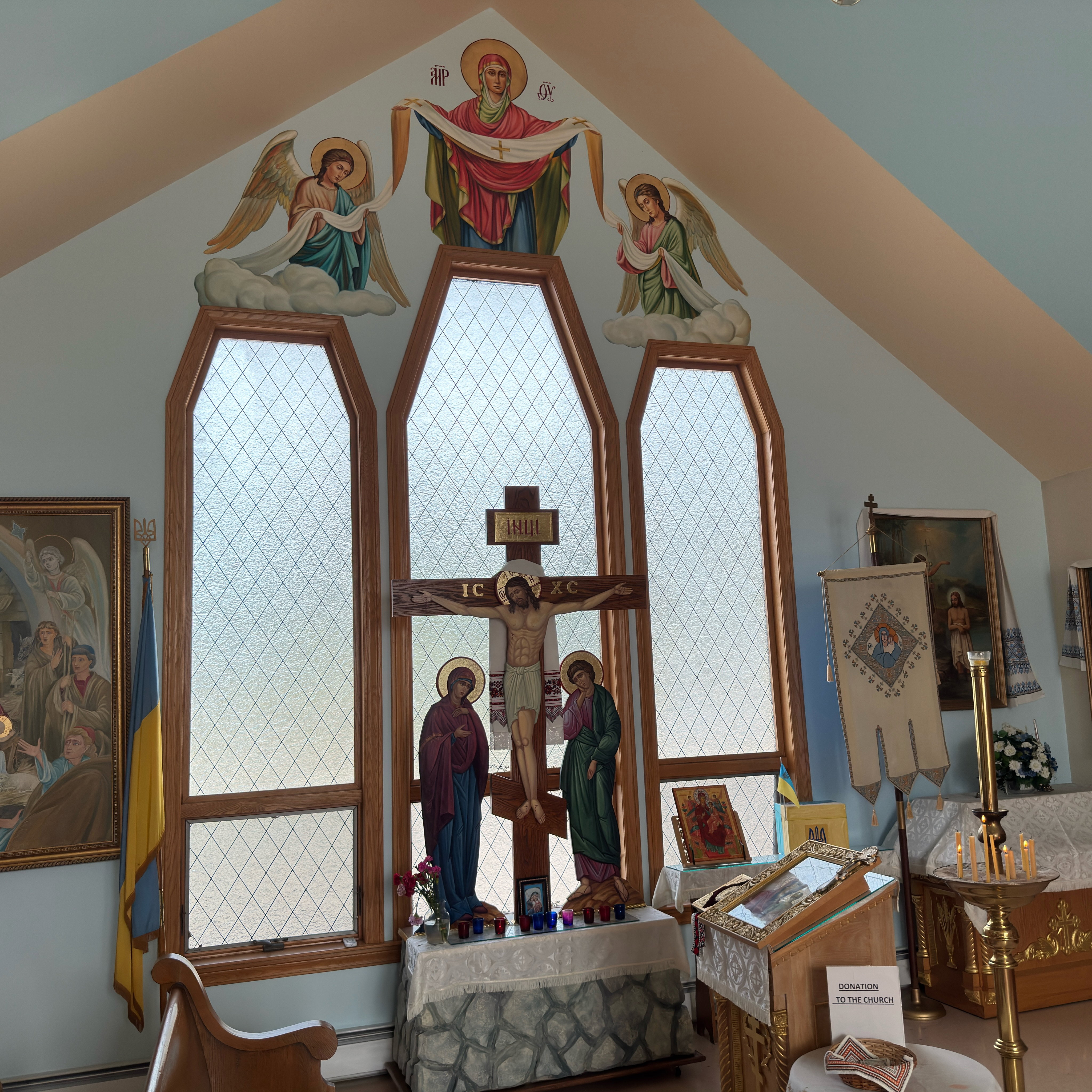 The east gable wall of Holy Trinity: above three tall leaded-glass windows, a painting of the Protection of the Theotokos, marked MP ΘY, shows the Mother of God in a red maphorion spreading a white veil marked with a cross, its ends held by two angels standing on clouds; beneath it the painted Crucifixion inscribed IC XC and INЦІ, draped with an embroidered rushnyk, with the Theotokos and St. John beneath the cross, above a Golgotha table of votive candles, carnations and a small icon; to the left the Nativity painting and a Ukrainian flag topped with a gold trident; to the right an analogion with an icon under glass, an icon of the Theotokos, an embroidered processional banner, a brass candle stand with beeswax tapers and a donation table