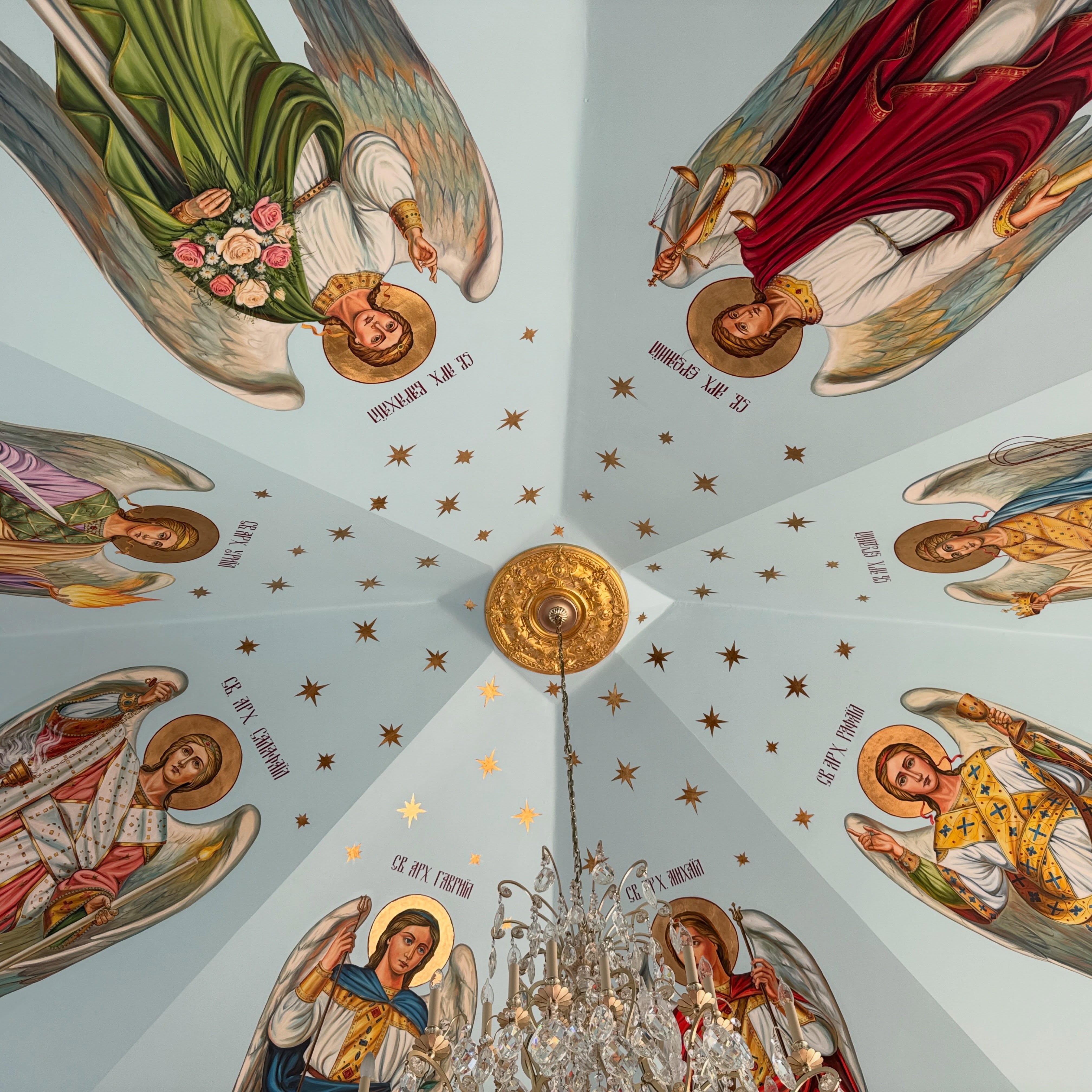 Close view of the archangel ceiling fresco at Holy Trinity: eight haloed archangels in green, red, lilac, blue, rose and gold vestments, each named in Church Slavonic (Cyrillic) lettering, around a gilded rosette, gold stars and a crystal chandelier