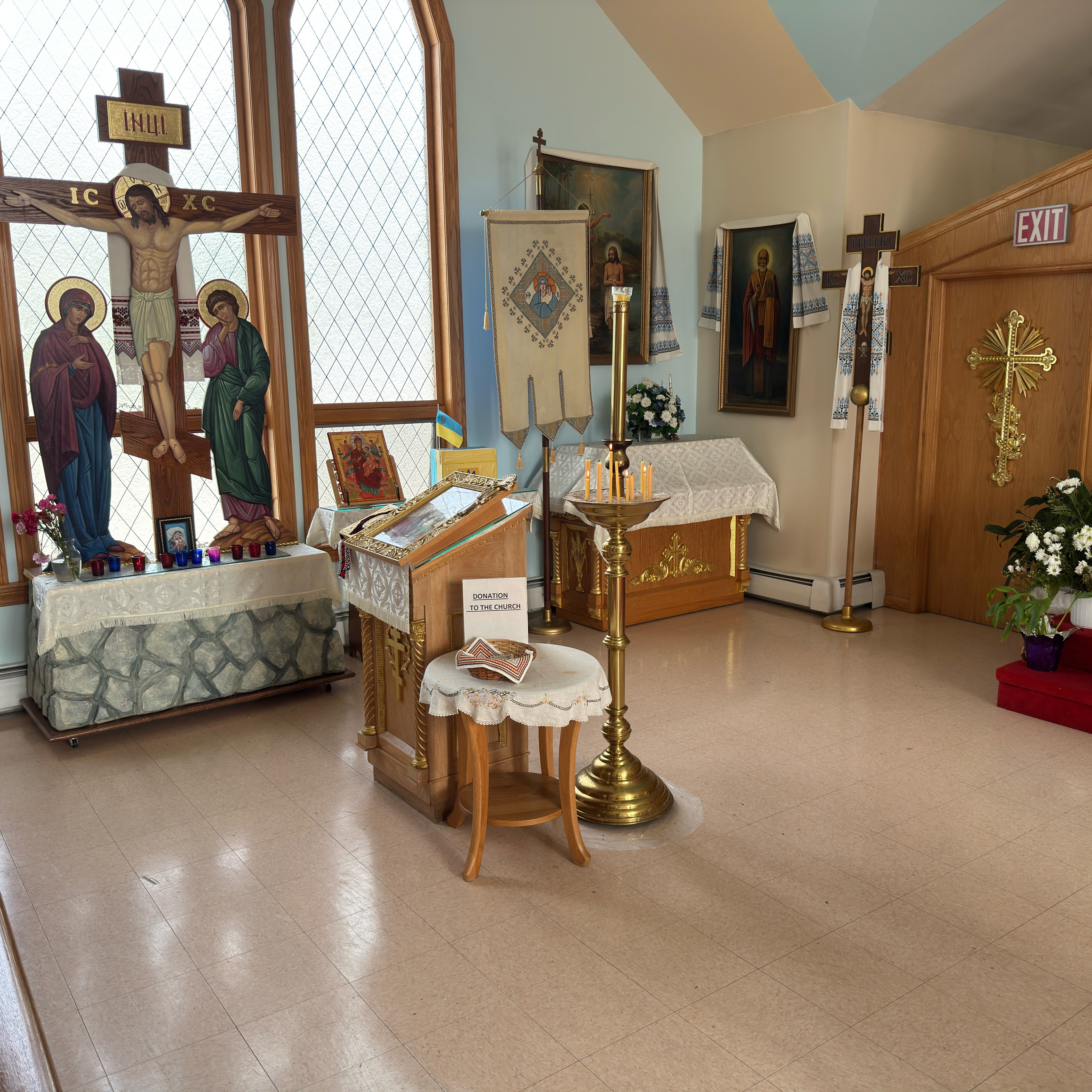 A sunlit corner of Holy Trinity: a large painted Crucifixion inscribed IC XC and INЦI, with the Theotokos and St. John standing beneath the cross before tall leaded-glass windows, above a Golgotha table of votive candles and carnations; beside it a carved analogion with a framed icon, a donation table, a brass candle stand with beeswax tapers, a processional banner of the Theotokos, an icon of St. Nicholas draped in an embroidered rushnyk, a processional cross and a gilded cross on the wooden sanctuary door