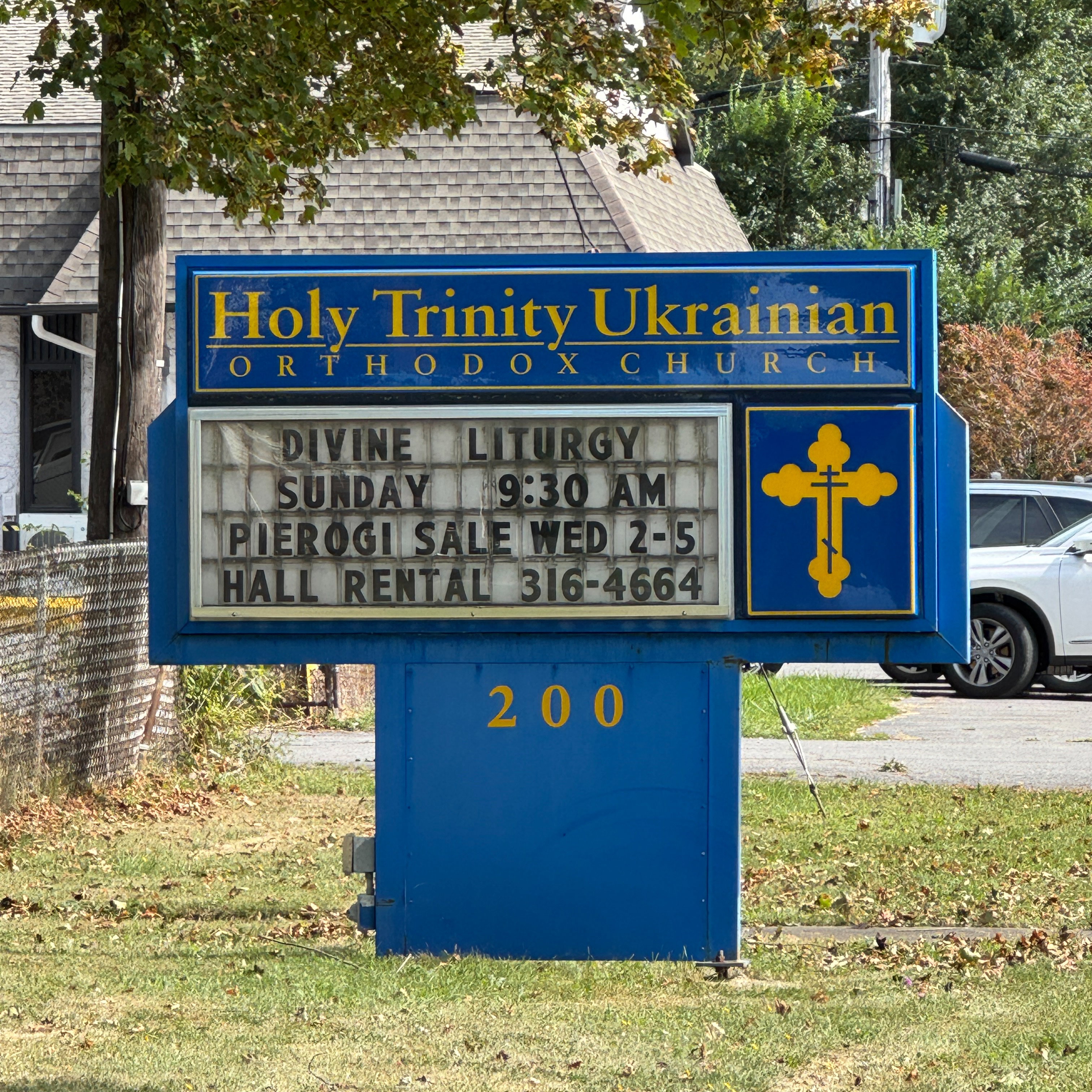The blue parish sign outside Holy Trinity Ukrainian Orthodox Church, lettered in gold with a gold three-bar Orthodox cross and the street number 200; its letter board reads Divine Liturgy Sunday 9:30 AM, Pierogi Sale Wed 2–5 and Hall Rental; behind it the church's shingled roof, green trees and the parking lot on a sunny day