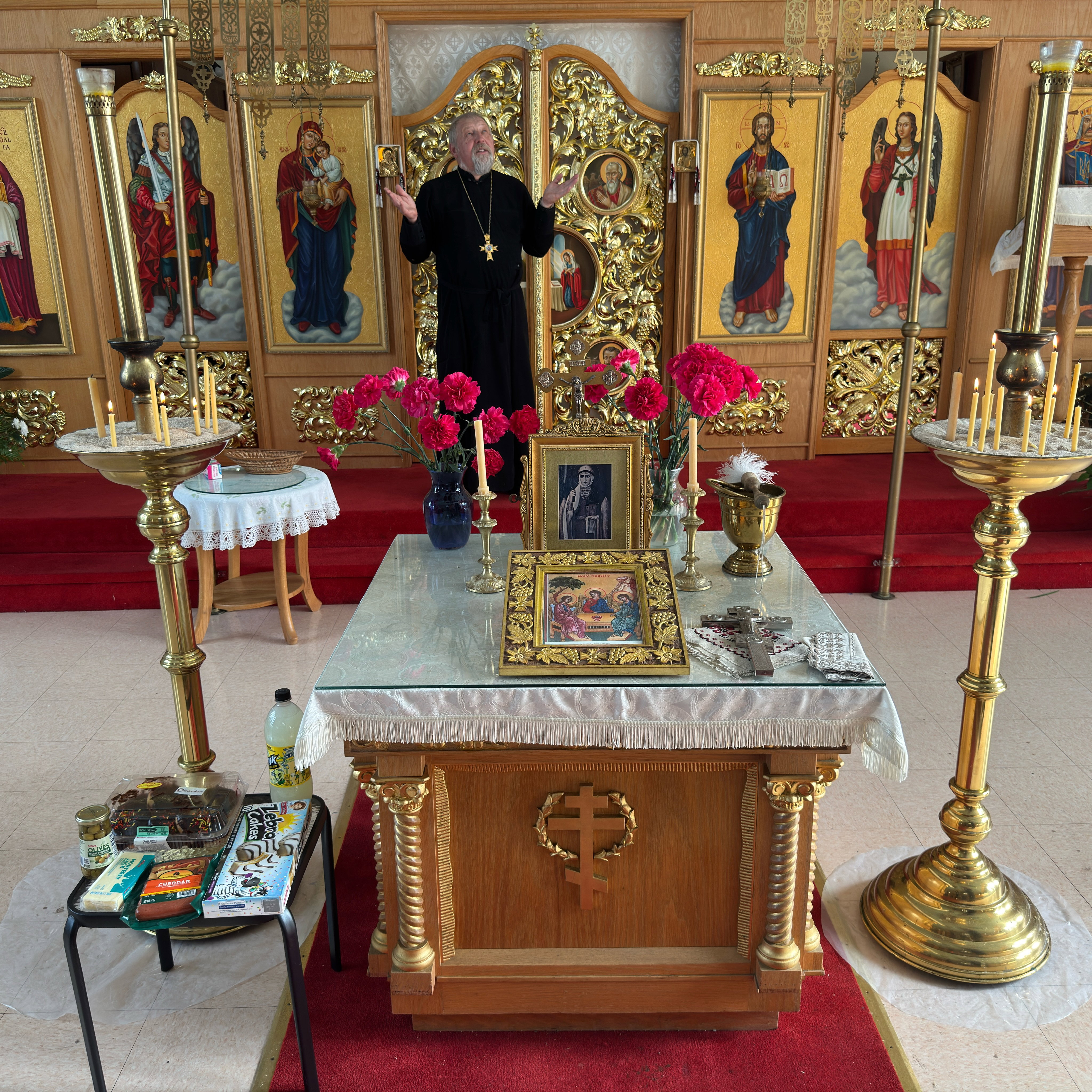 A priest in a black cassock and pectoral cross raises his hands in prayer before the gilded Royal Doors of the iconostasis at Holy Trinity, flanked by icons of the archangels, the Theotokos and Christ; in front, the altar table holds a framed Holy Trinity icon, a blessing cross, beeswax candles and red carnations between two tall brass candle stands