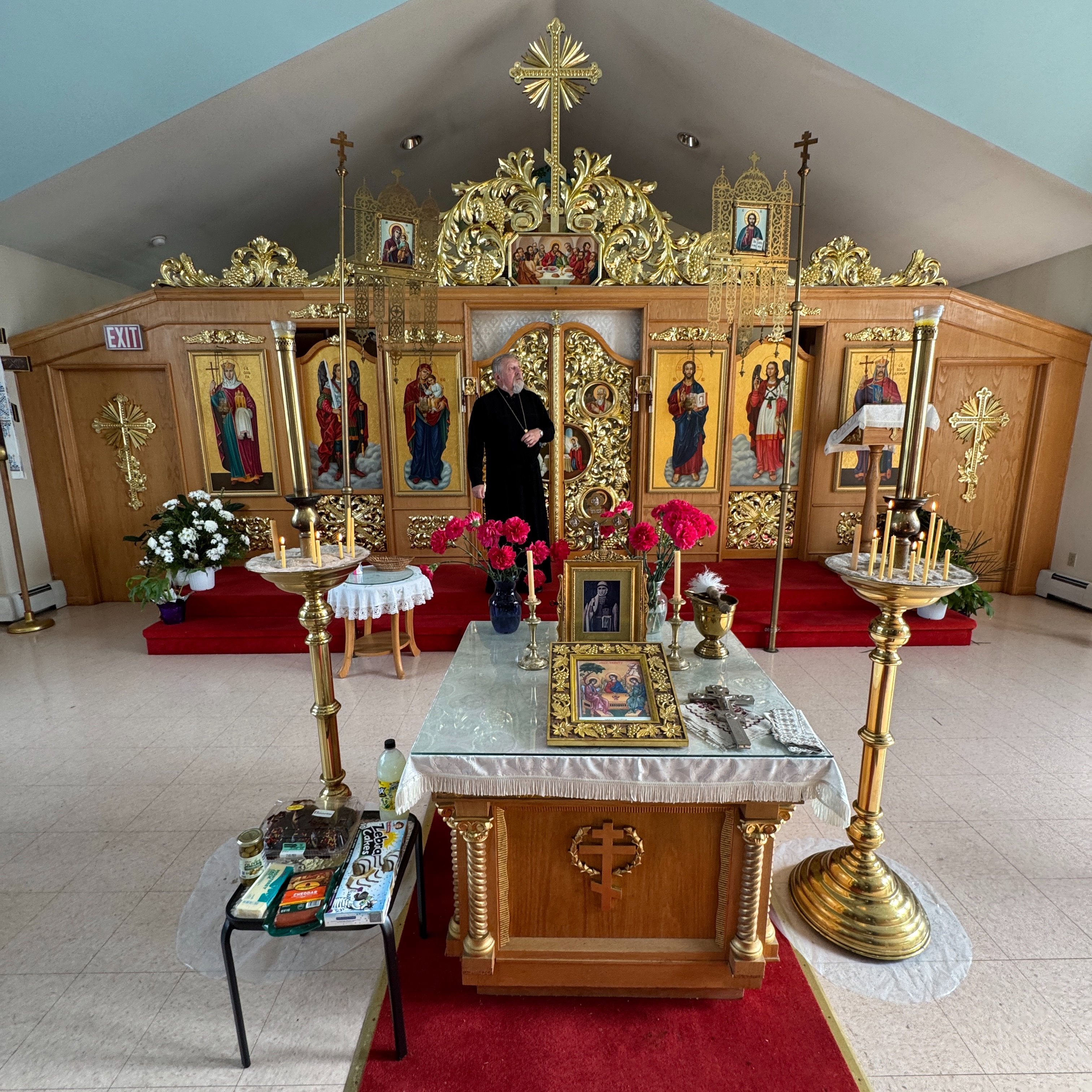 Wide view of the full iconostasis at Holy Trinity: a priest in a black cassock and pectoral cross stands at the open gilded Royal Doors, between icons of St. Olga, Archangel Michael, the Theotokos, Christ, Archangel Gabriel and St. Volodymyr, beneath the Last Supper, carved gold vine scrollwork and a radiant gold cross; in the foreground a table draped in white holds a gilt-framed Holy Trinity icon, a blessing cross, a portrait icon, beeswax candles and red carnations between two tall brass candle stands, beside a small table of offerings on a red carpet