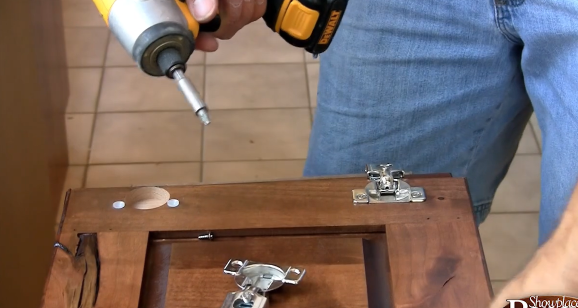 Homeowner kneeling on a drop cloth, measuring a new white cabinet end panel against a run of cream shaker cabinets with a speed square and clamps nearby