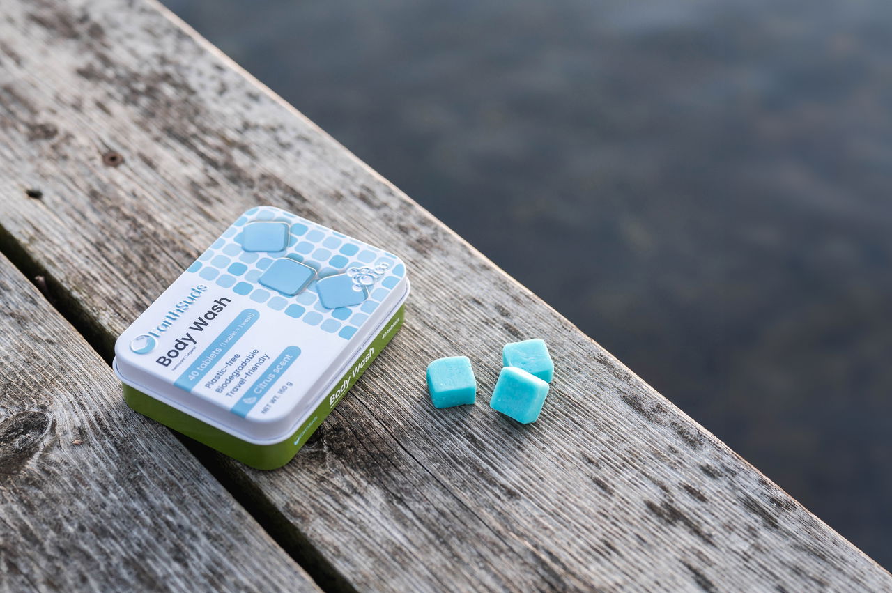 EarthSuds Body Wash tin sits on a weathered wooden surface by the water, with blue shower tablets placed nearby — evoking outdoor adventure or eco-travel.
