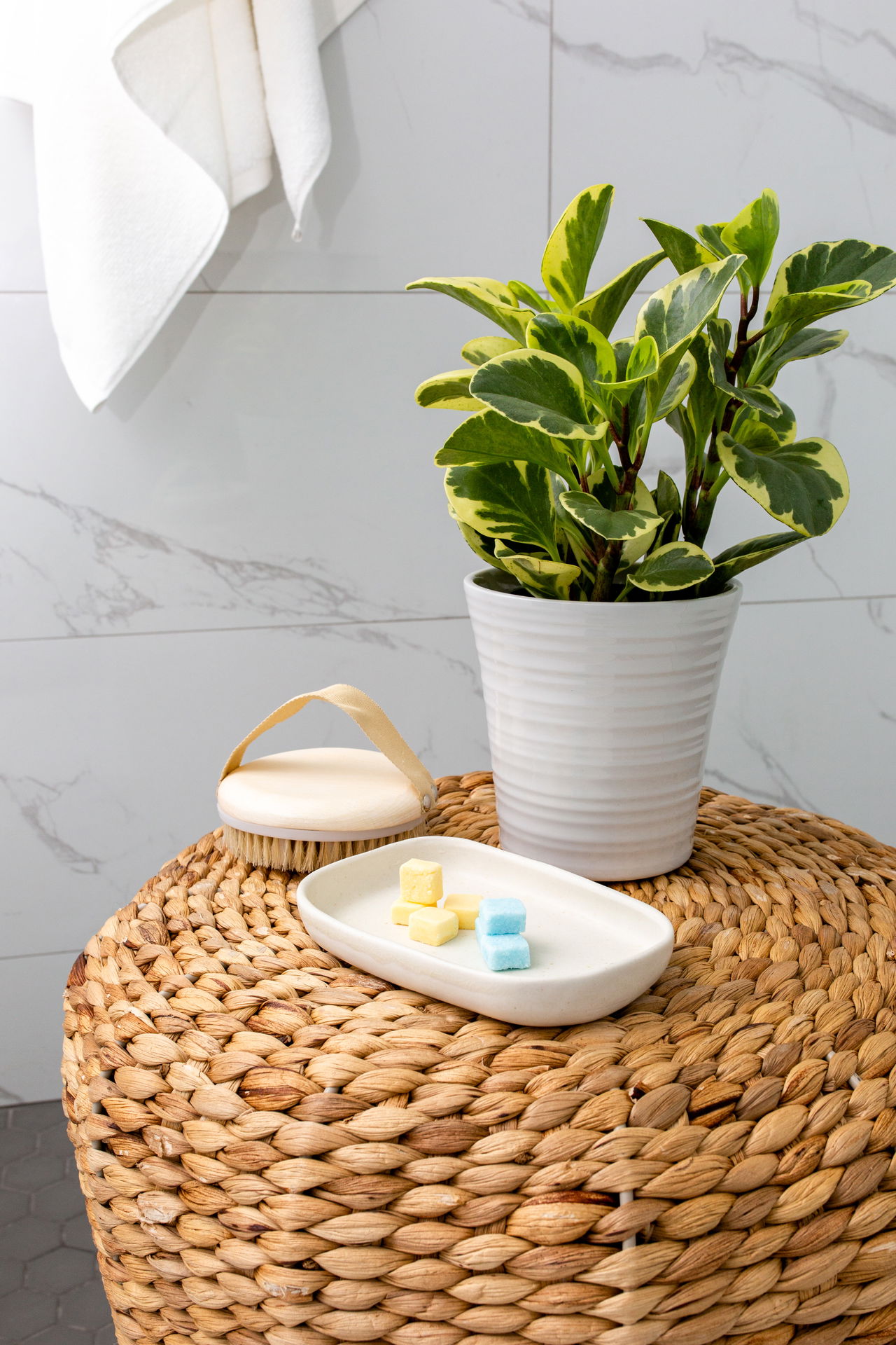 EarthSuds shower tablets in a white soap dish sit on a woven bathroom stool beside a potted plant, with a white towel hanging in the background — a clean, minimalist setup highlighting natural and eco-friendly self-care.