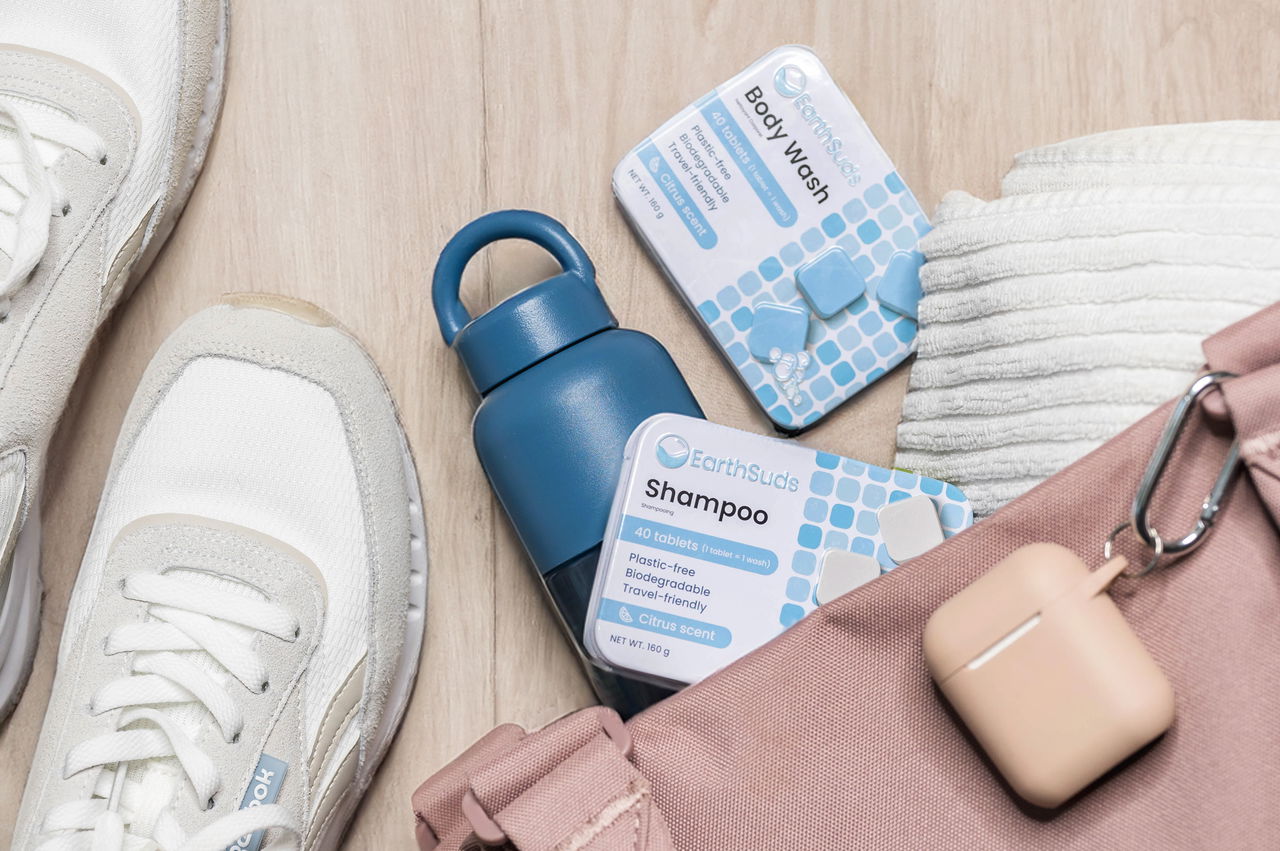 EarthSuds Shampoo and Body Wash boxes rest on a wooden floor next to sneakers, a water bottle, and workout gear, highlighting gym or travel use.