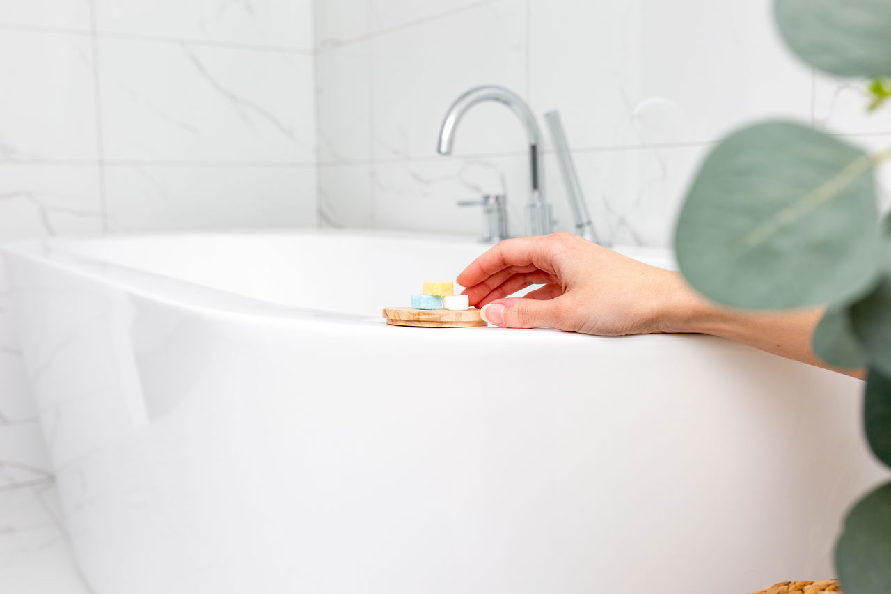 A hand reaches for solid shampoo tablets resting on a small wooden dish beside a modern bathtub. A sleek chrome faucet and white tiled wall are in the background, with a eucalyptus plant in the foreground. Text overlay reads: “Compact, travel-friendly shower tablets. Solid shampoo tablets that eliminate plastic and mess – perfect for travel, everyday use.”