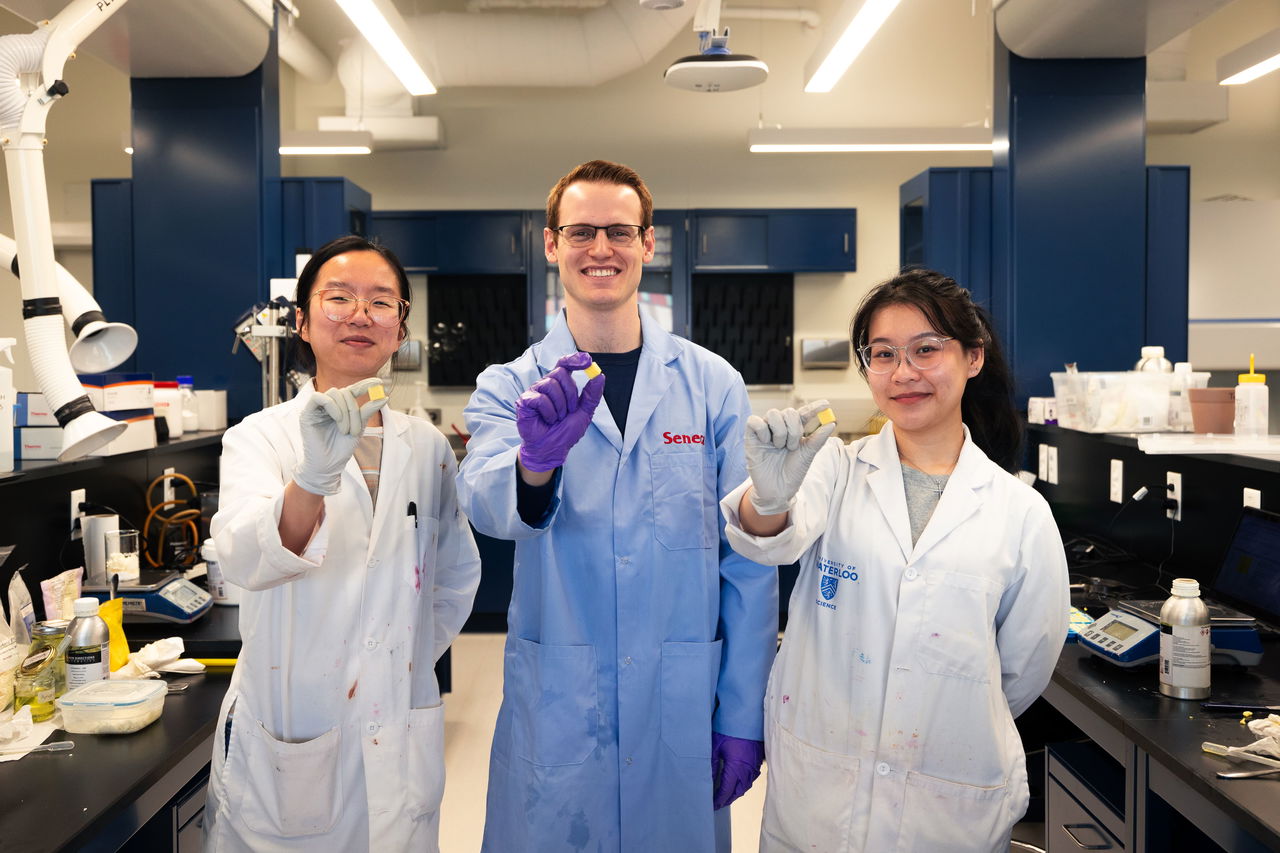 Three lab researchers wearing branded lab coats and gloves stand in a modern laboratory, smiling and holding up EarthSuds shower tablets, showcasing innovation and product development in a scientific setting.