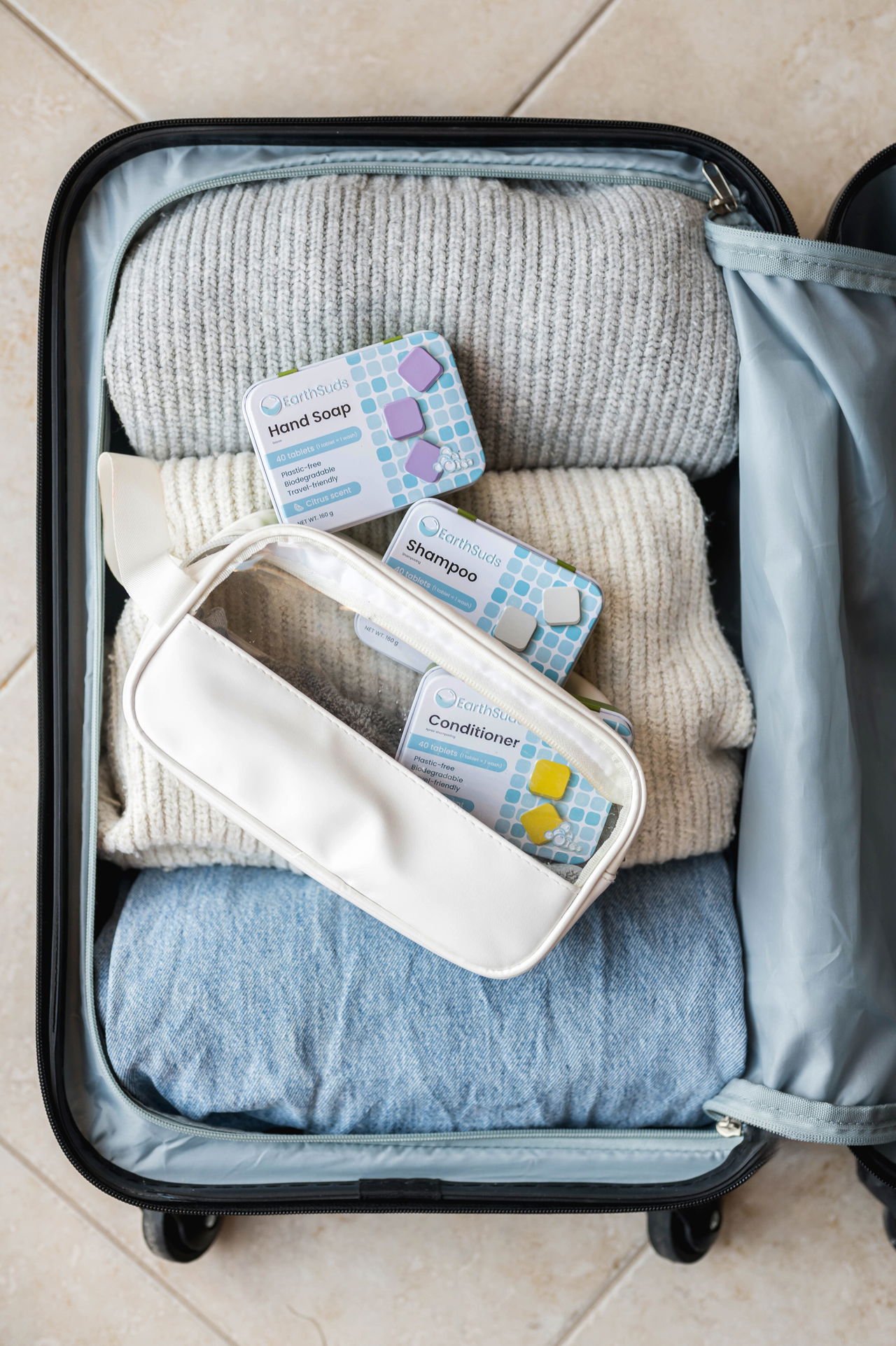 An open suitcase neatly packed with folded clothing and a white toiletry bag. On top are three boxes of EarthSuds solid shower tablets labeled Hand Soap, Shampoo, and Conditioner, showcasing a compact and travel-friendly format.