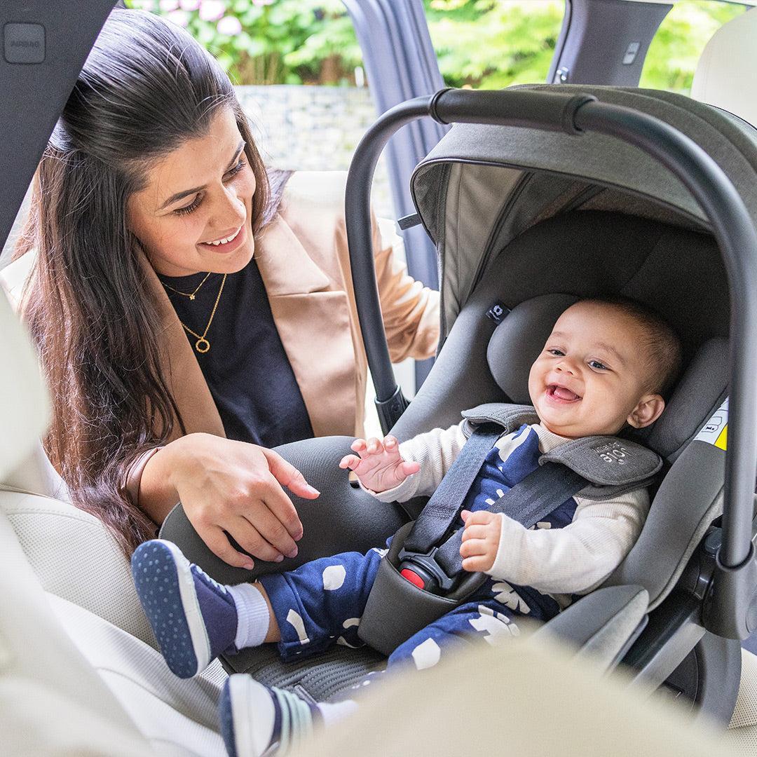 Mother smiling at her laughing baby buckled into a grey Joie infant car seat in the back of a car