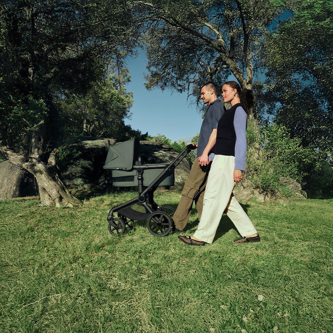 Couple walking hand in hand across sunny parkland pushing a forest green Bugaboo pushchair with carrycot