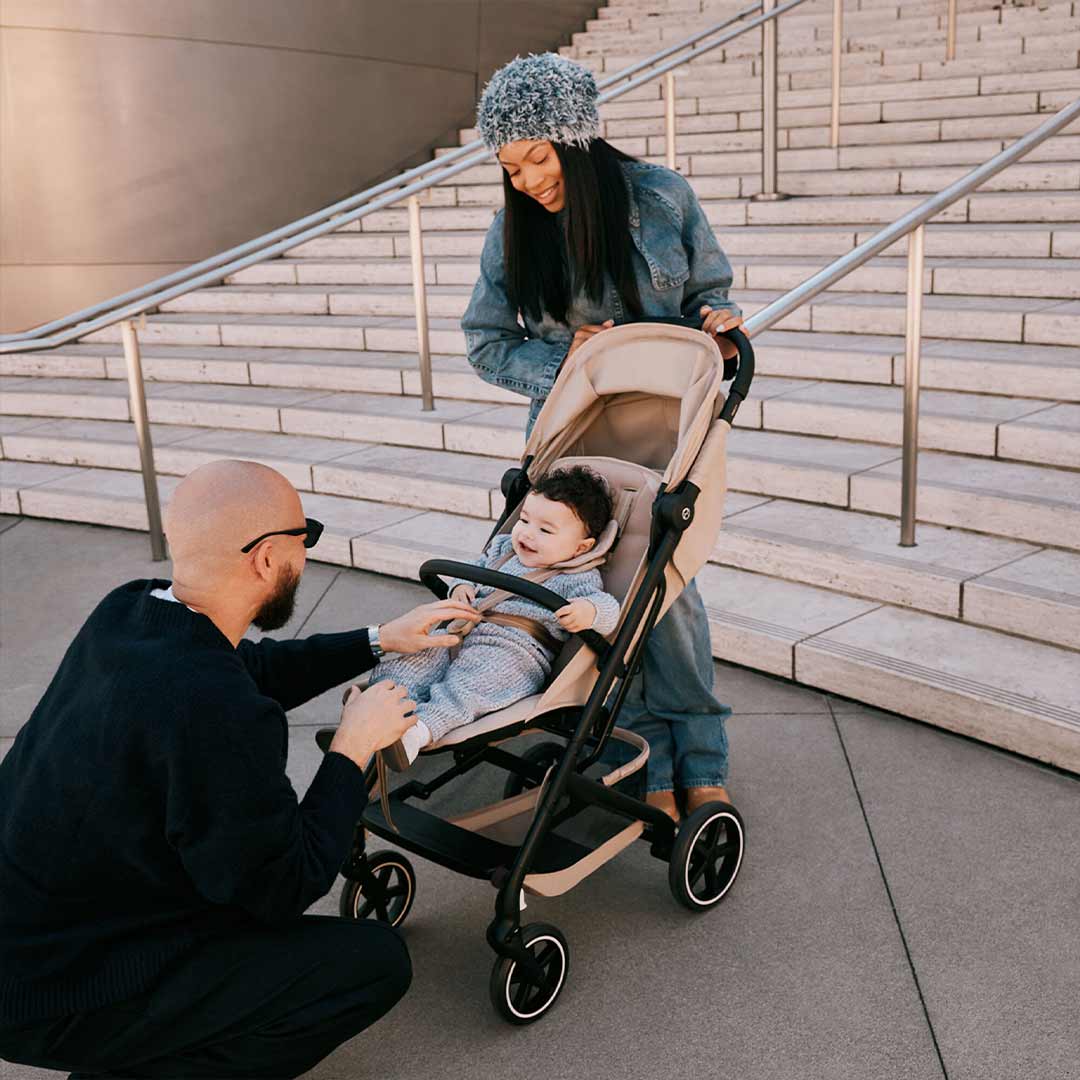 Smiling parents with their baby in a beige CYBEX Beezy stroller beside sunlit stone steps