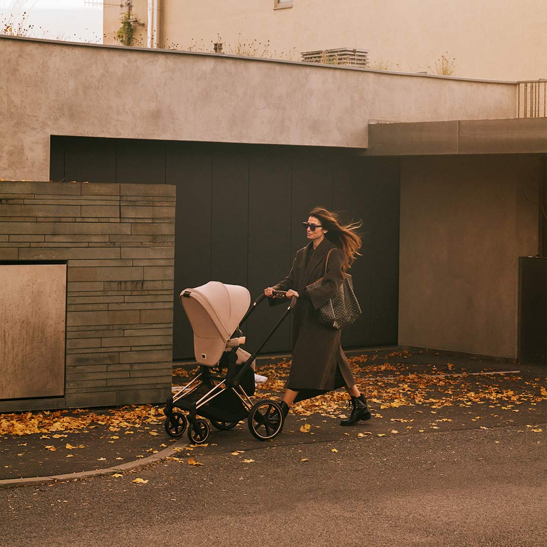 Woman in a long coat pushing a stroller past autumn leaves