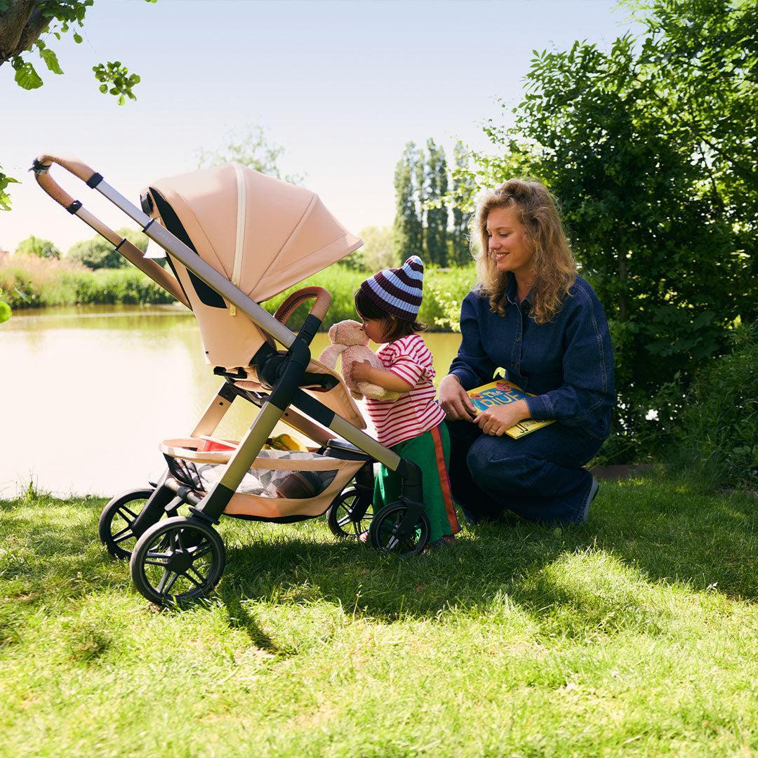 Smiling mother kneeling on the grass by a lake beside a peach Joolz pushchair as her toddler hugs a teddy